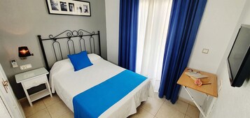 Una cama bien hecha con un cojín azul decorativo, una mesita de noche con una lámpara, una pequeña mesita de madera con un libro y una rosa, y un televisor montado en la pared.