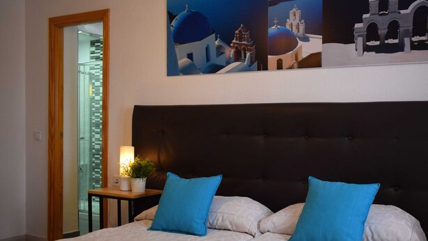Una habitación de hotel con una cama grande, dos almohadas azules, una mesita de noche con una lámpara y una planta, y una pared con un cuadro de un edificio.