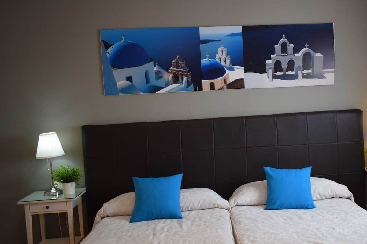 A hotel room with a large bed, two blue pillows, a nightstand with a lamp, and a wall-mounted painting of a coastal scene.
