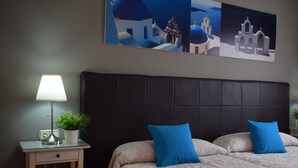 Habitación de hotel con cama, mesita de noche con lámpara, planta en maceta y un cuadro de una escena costera.