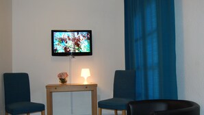 Una habitación con televisión montada en la pared, dos sillas, una mesa con lámpara y una ventana con cortinas azules.