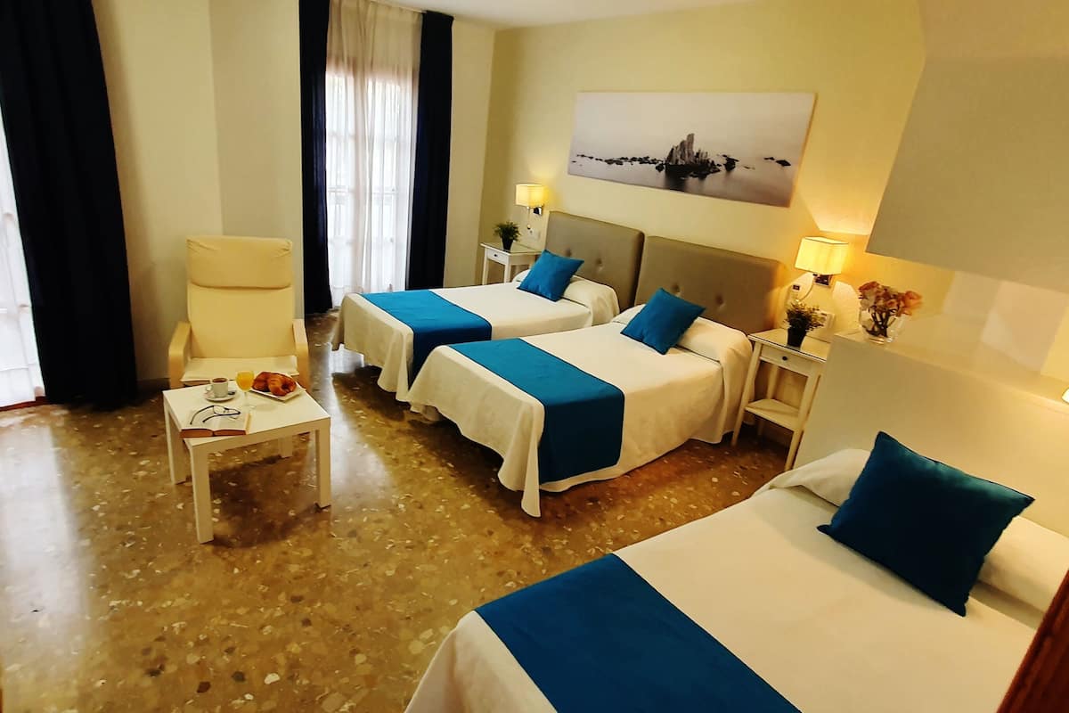 A hotel room with two beds, a small table with a book and a plate of fruit, a chair, and a nightstand with a lamp.