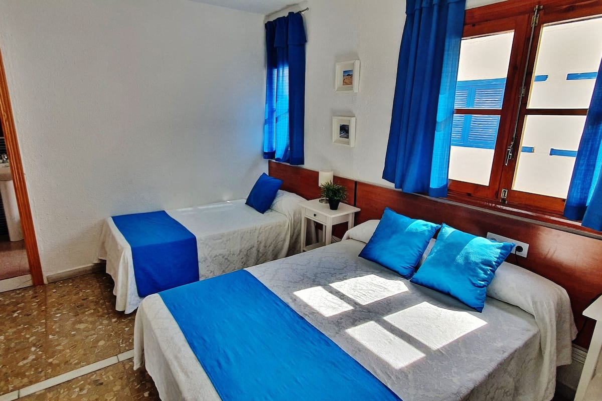 A hotel room with two beds, a small table, a nightstand, a window with blue curtains, and a door leading to a bathroom.