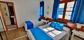 Habitación de hotel con dos camas, una mesita pequeña, una mesita de noche, una ventana con cortinas azules y una puerta que da a un baño.