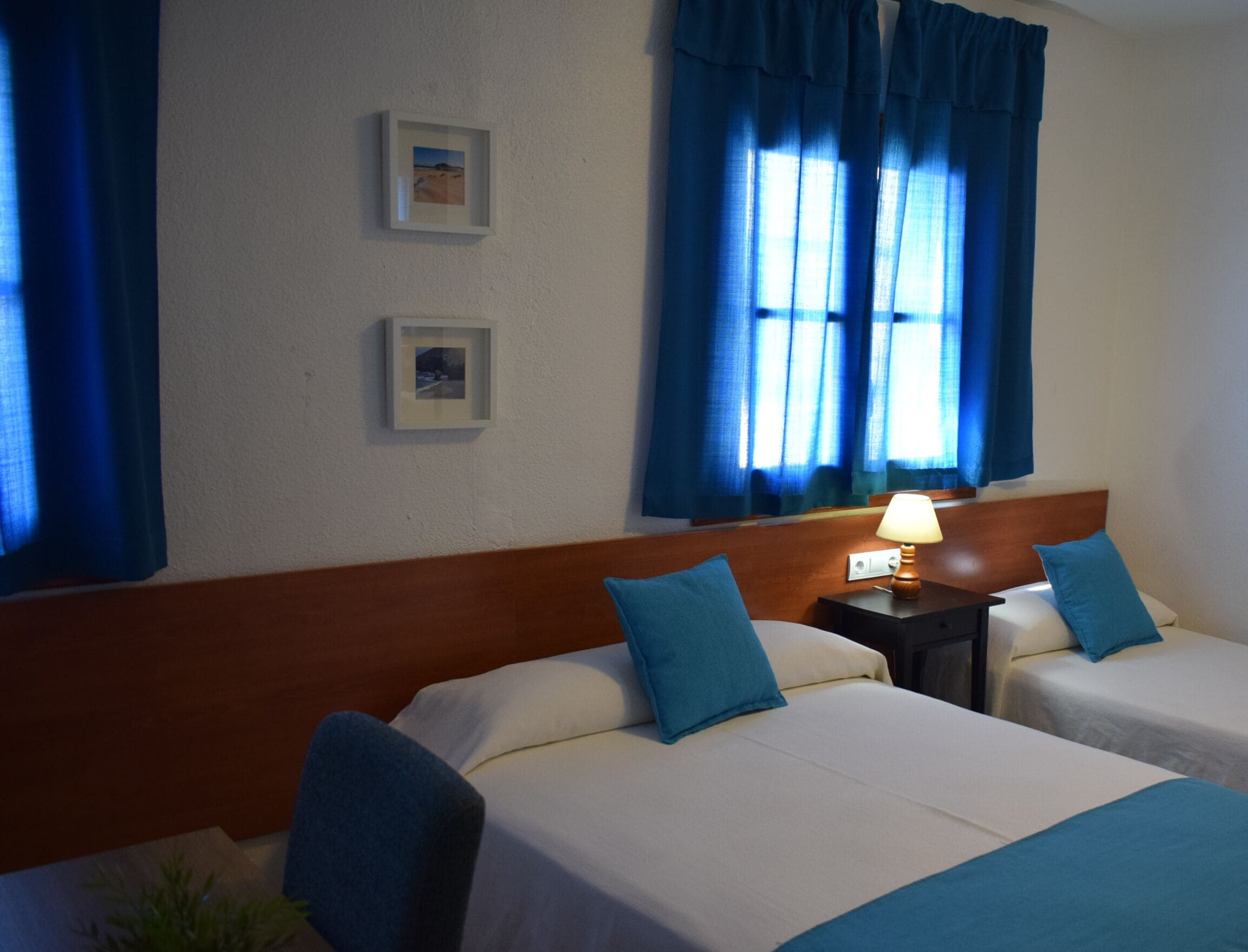 A hotel room with two beds, a nightstand with a lamp, and two framed pictures on the wall.