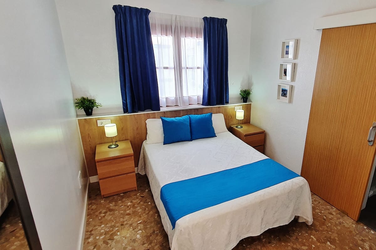 A hotel room with a bed, bedside tables, a nightstand, a window with curtains, a wooden door, and a wall-mounted picture.
