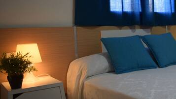 Una cama bien hecha con colcha blanca y azul, una lámpara de noche y una planta en maceta sobre una mesita blanca.