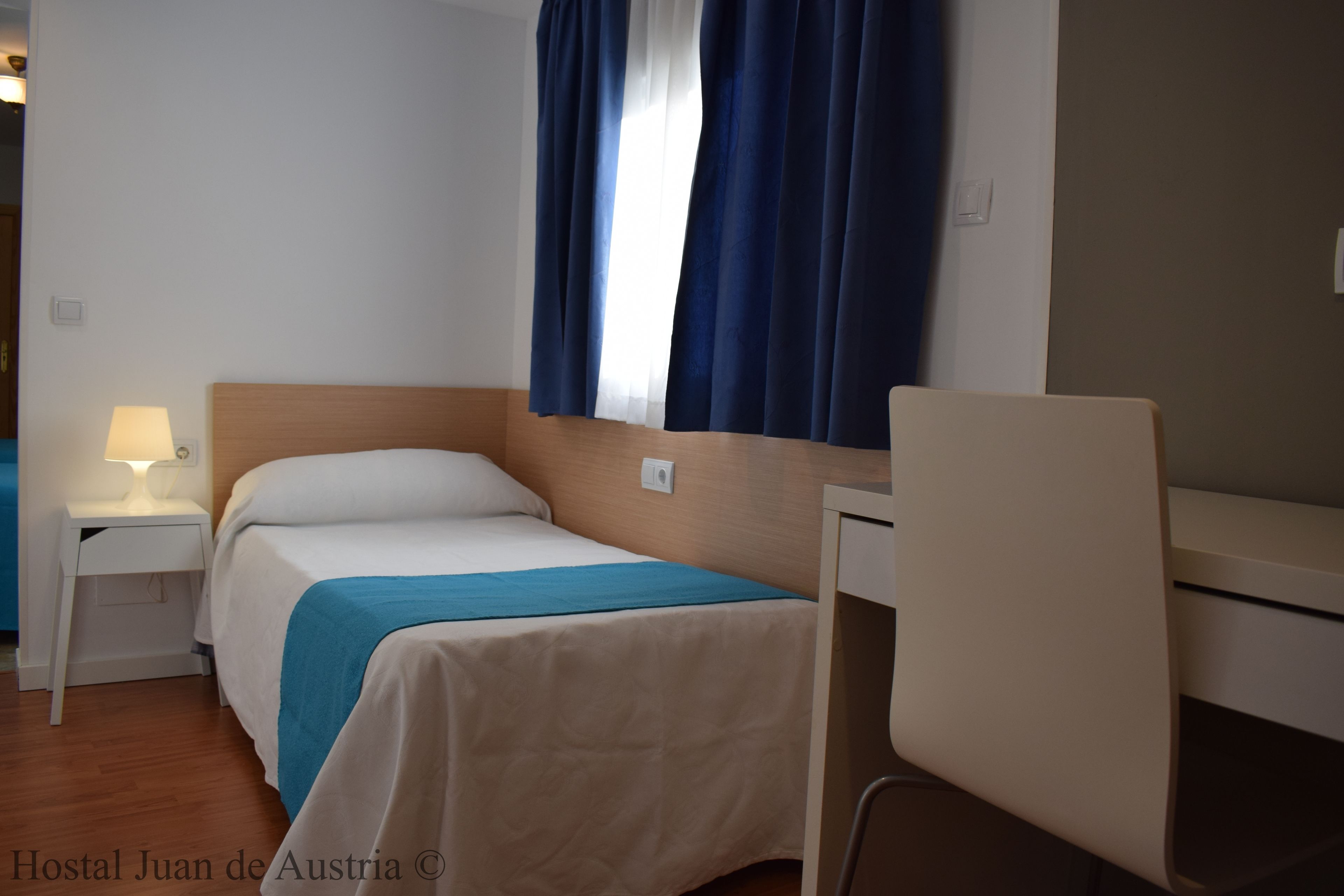 Habitación de hotel con cama, escritorio, silla, mesita de noche con lámpara y ventana con cortinas.