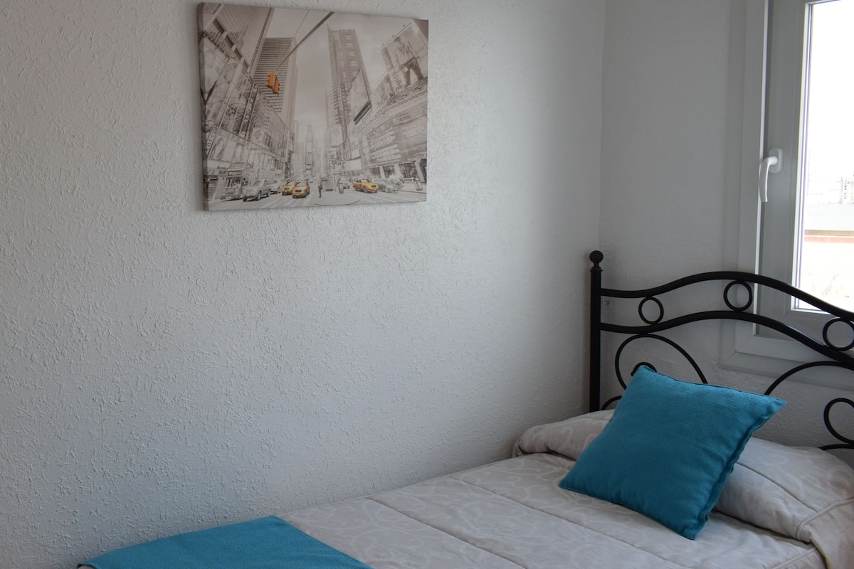 A single bed with a blue pillow and a blue blanket, a black metal headboard, and a wall-mounted painting of a cityscape.