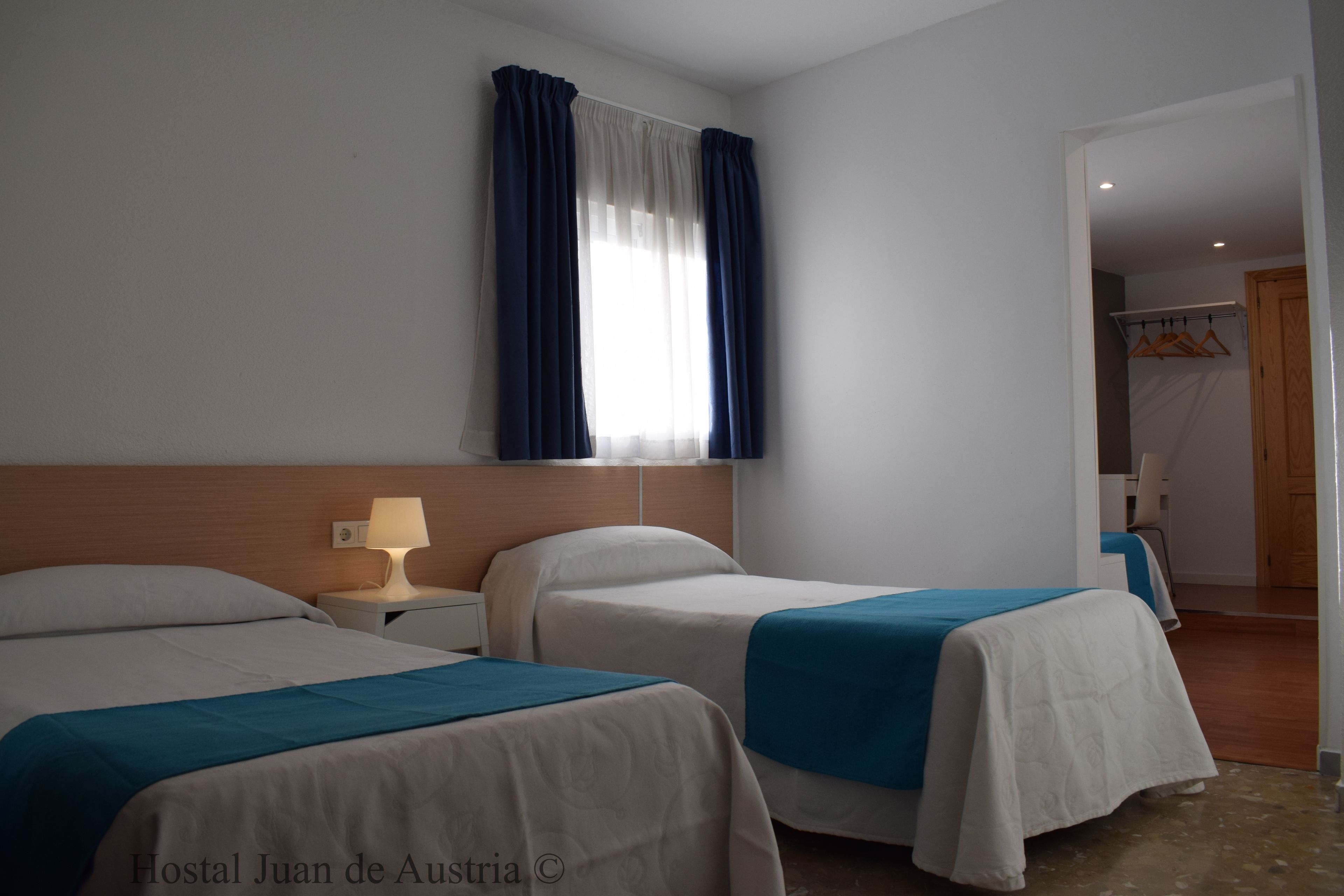 Habitación de hotel con dos camas, una mesita de noche con lámpara, una ventana con cortinas y vistas a otra habitación con perchero.