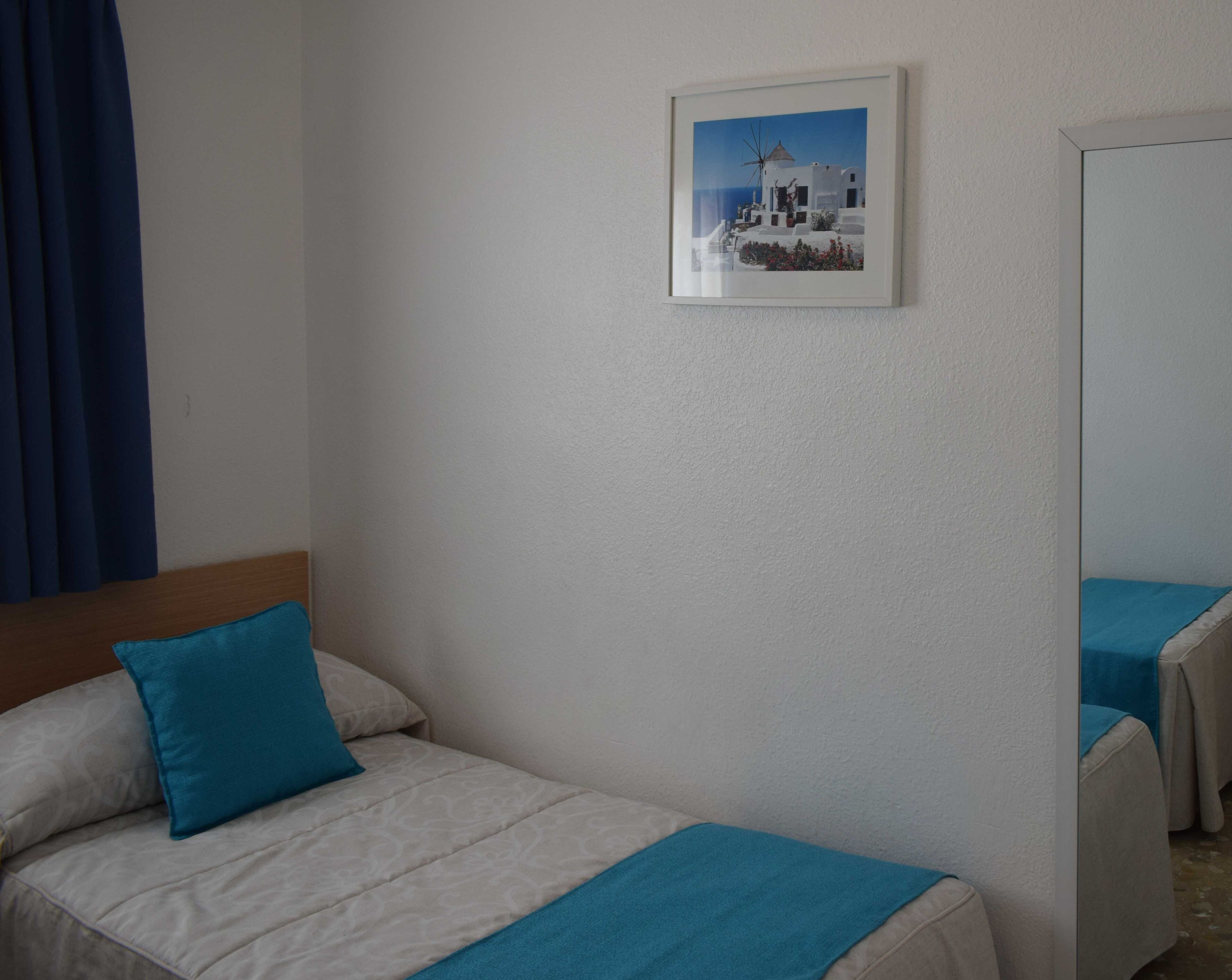 A hotel room with a bed, a blue pillow, a framed picture of a coastal scene, and a mirror reflecting part of the room.