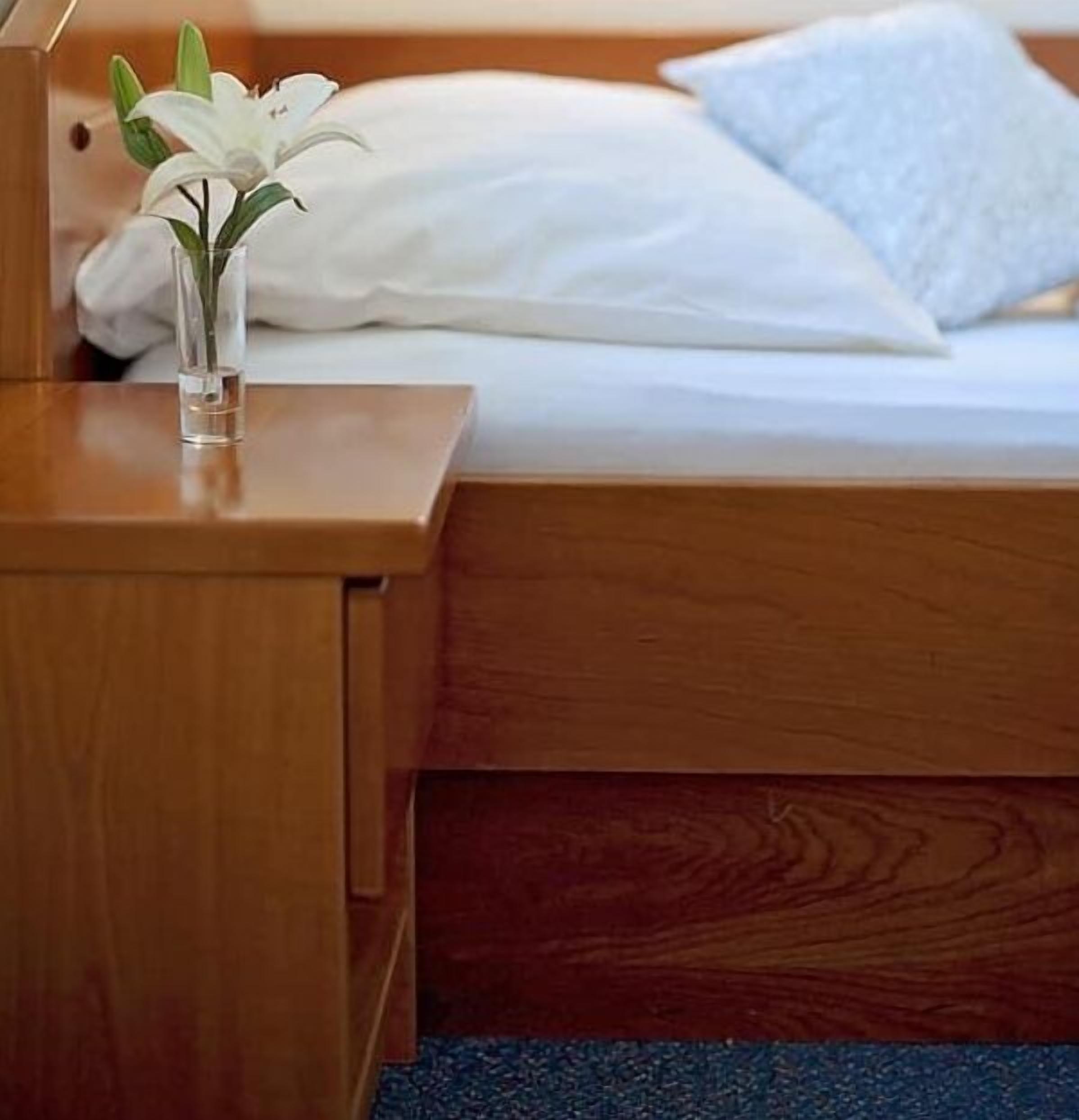 A neatly made bed with white linens and a single white pillow. A clear glass vase with a white flower is placed on a wooden bedside table.
