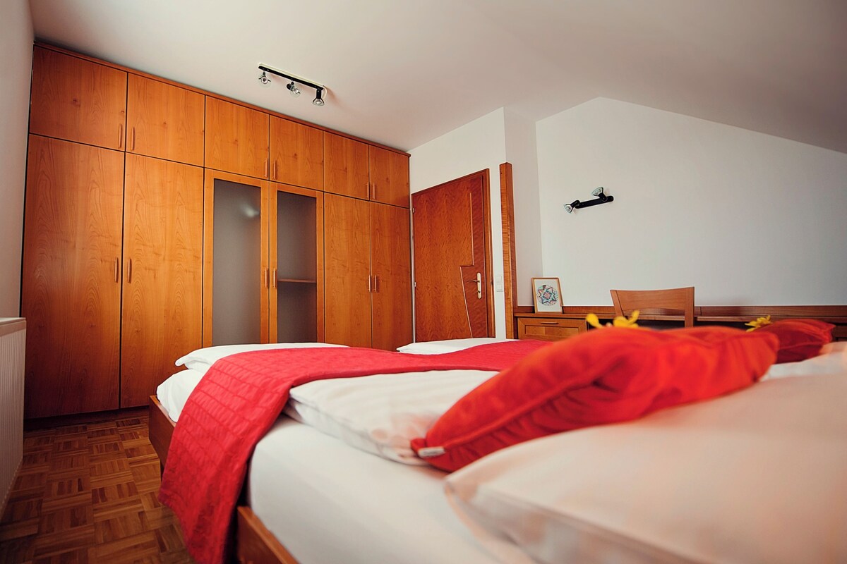 A bedroom with a wooden wardrobe, a bed with white bedding and a red blanket, and a wooden nightstand with a picture frame.