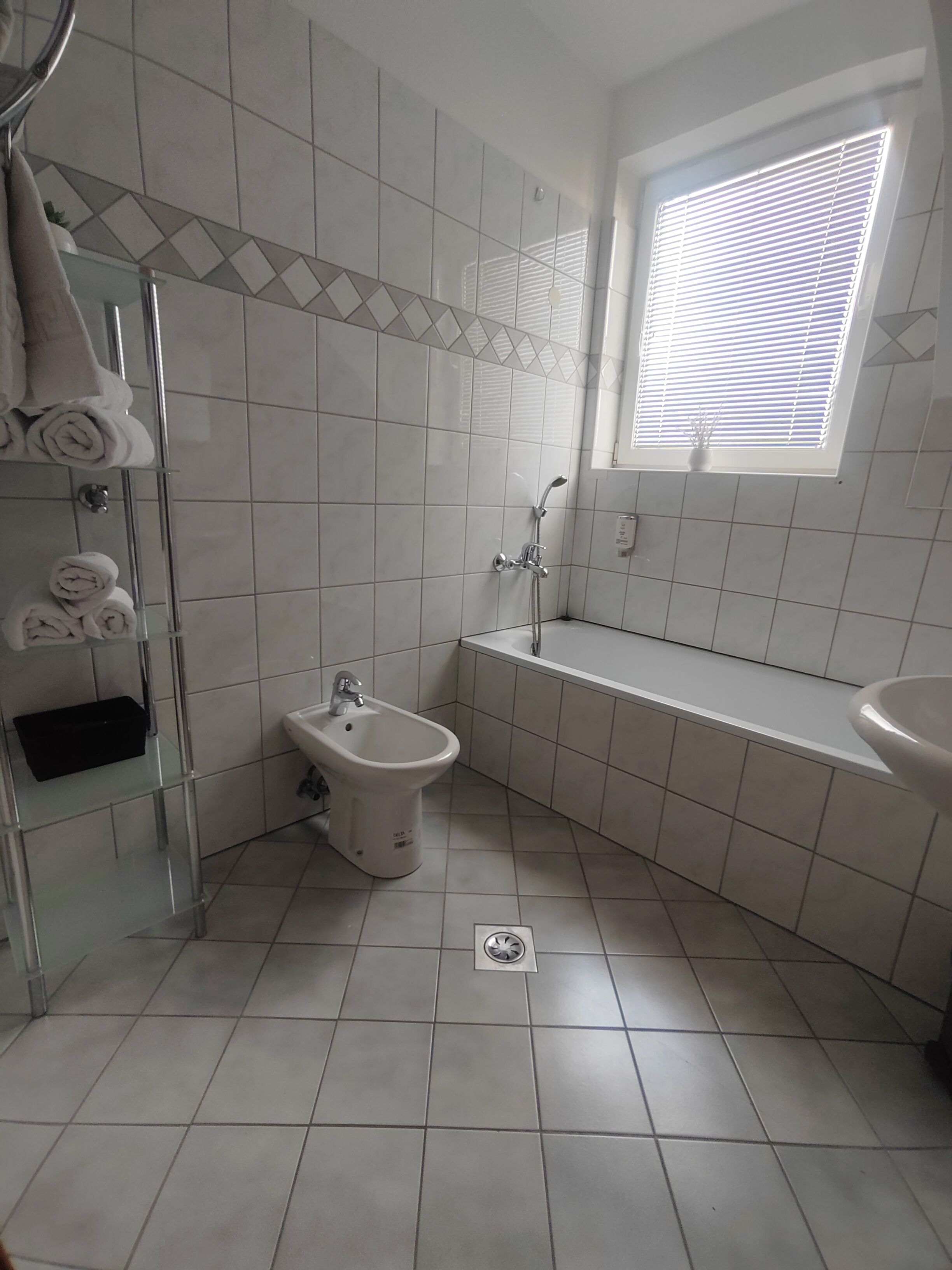 A bathroom with a bidet, a glass shower enclosure, a sink, and a window with blinds.