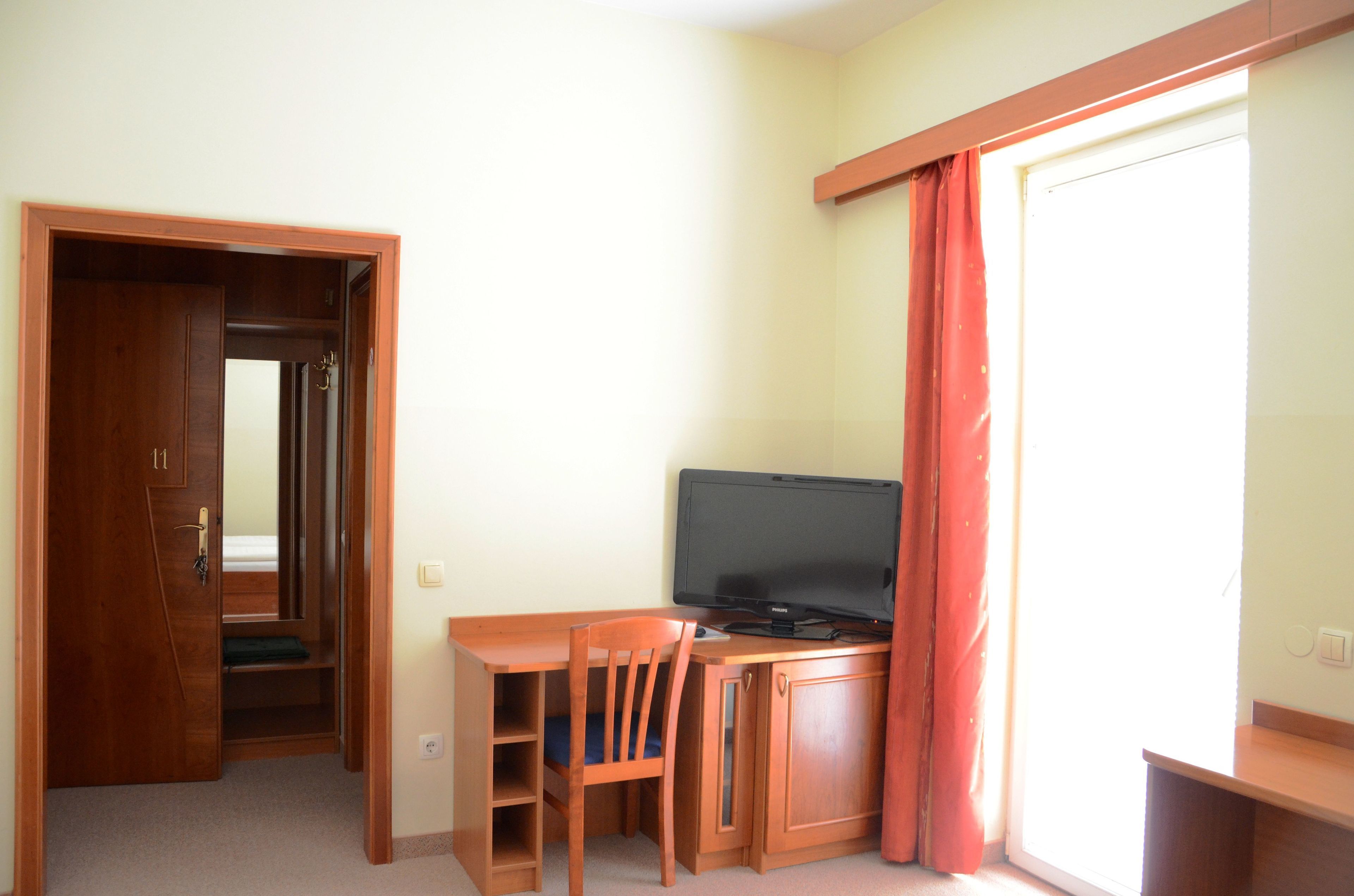 A compact room with a wooden desk, chair, and a television. There's a door leading to a closet and a window with curtains.