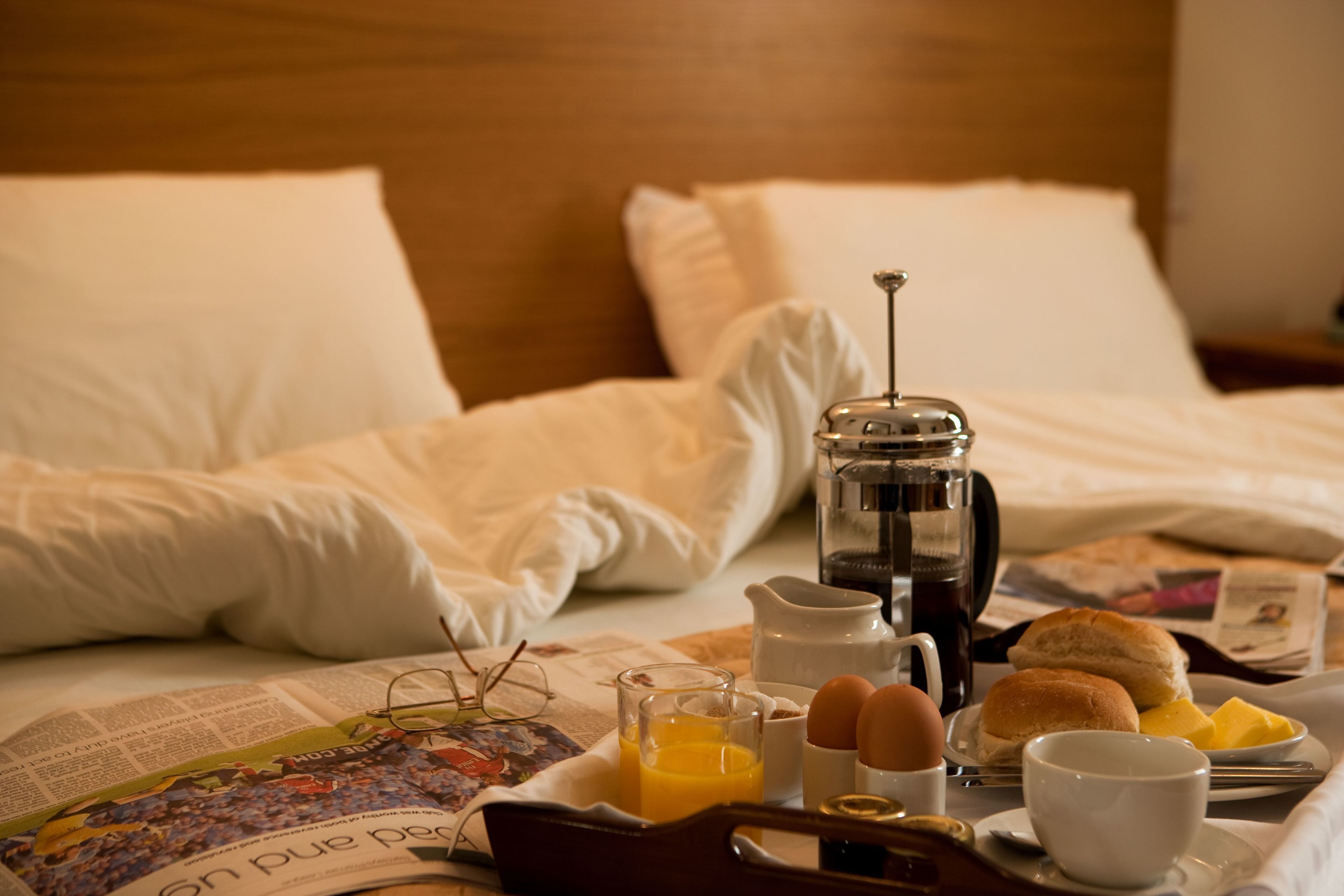 Un vassoio per la colazione ordinato su un letto, con un giornale, una caffettiera, uova e succo d'arancia.
