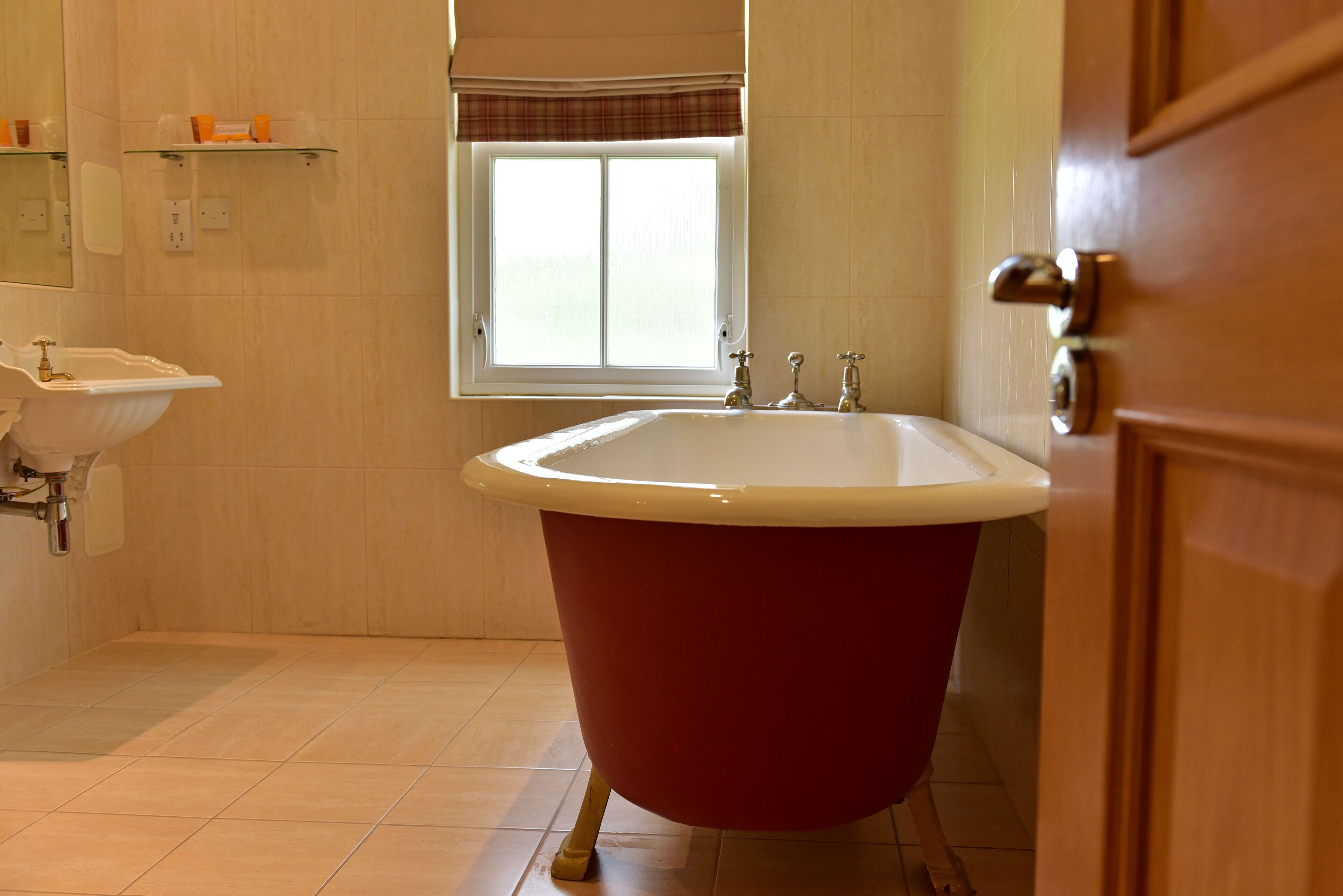 Un baño con una bañera roja de patas de león, un lavamanos blanco y una ventana con persianas.