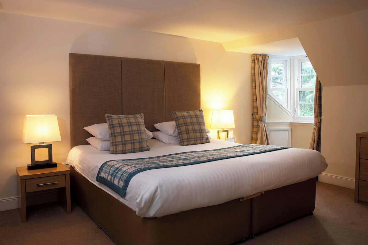 A neatly made bed with a headboard, two bedside tables with lamps, a window with curtains, and a wooden dresser.
