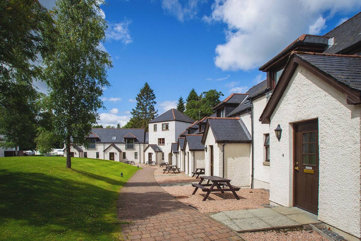 A row of white buildings with grey roofs, each with a door and windows, and a paved walkway with picnic tables.