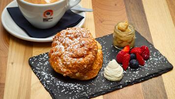 A dessert plate with a pastry, whipped cream, and berries on a slate board, a cup of coffee, and a jar of cream on a wooden table.