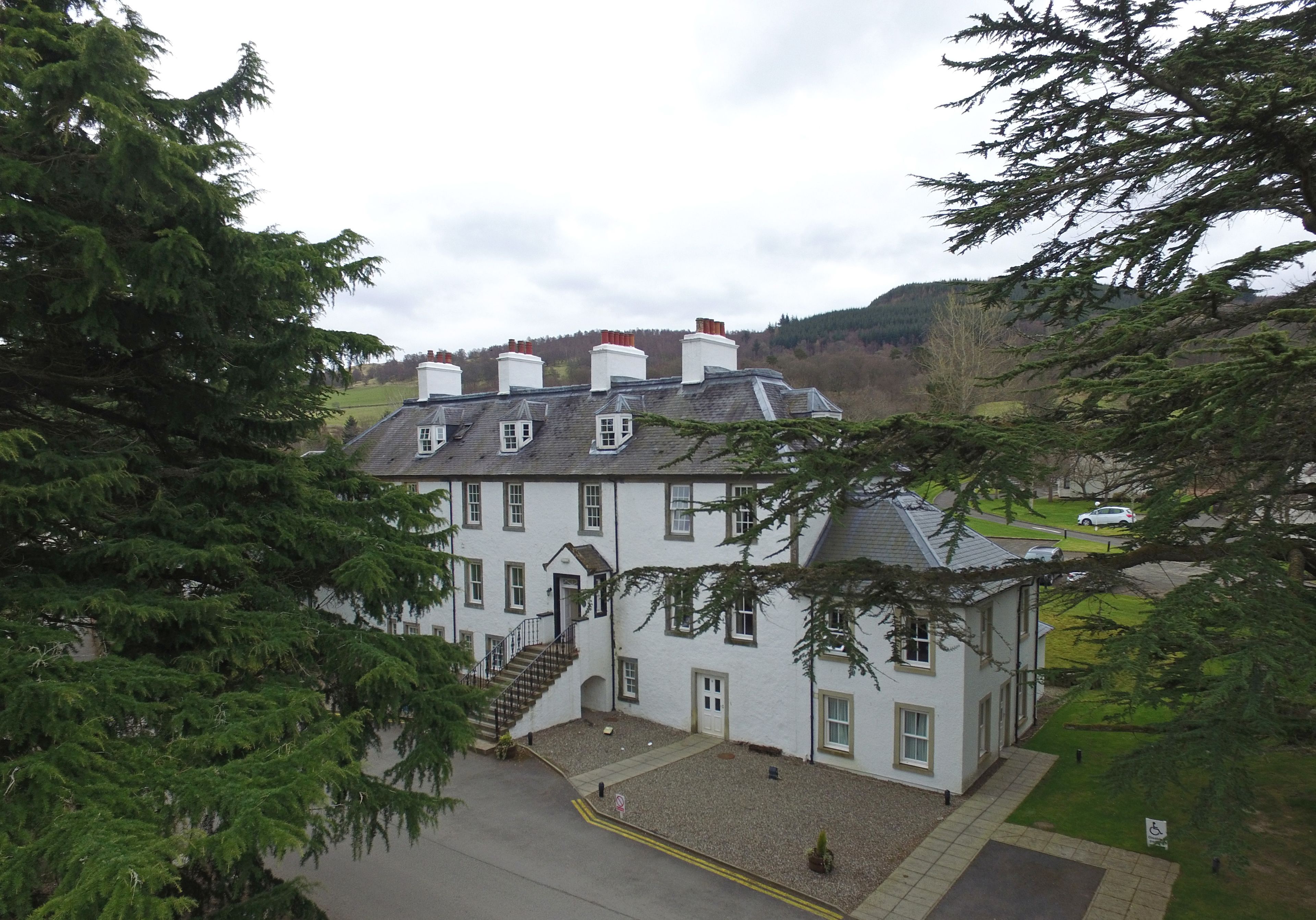 A large, white building with multiple windows and a central staircase, surrounded by trees and a hilly landscape.