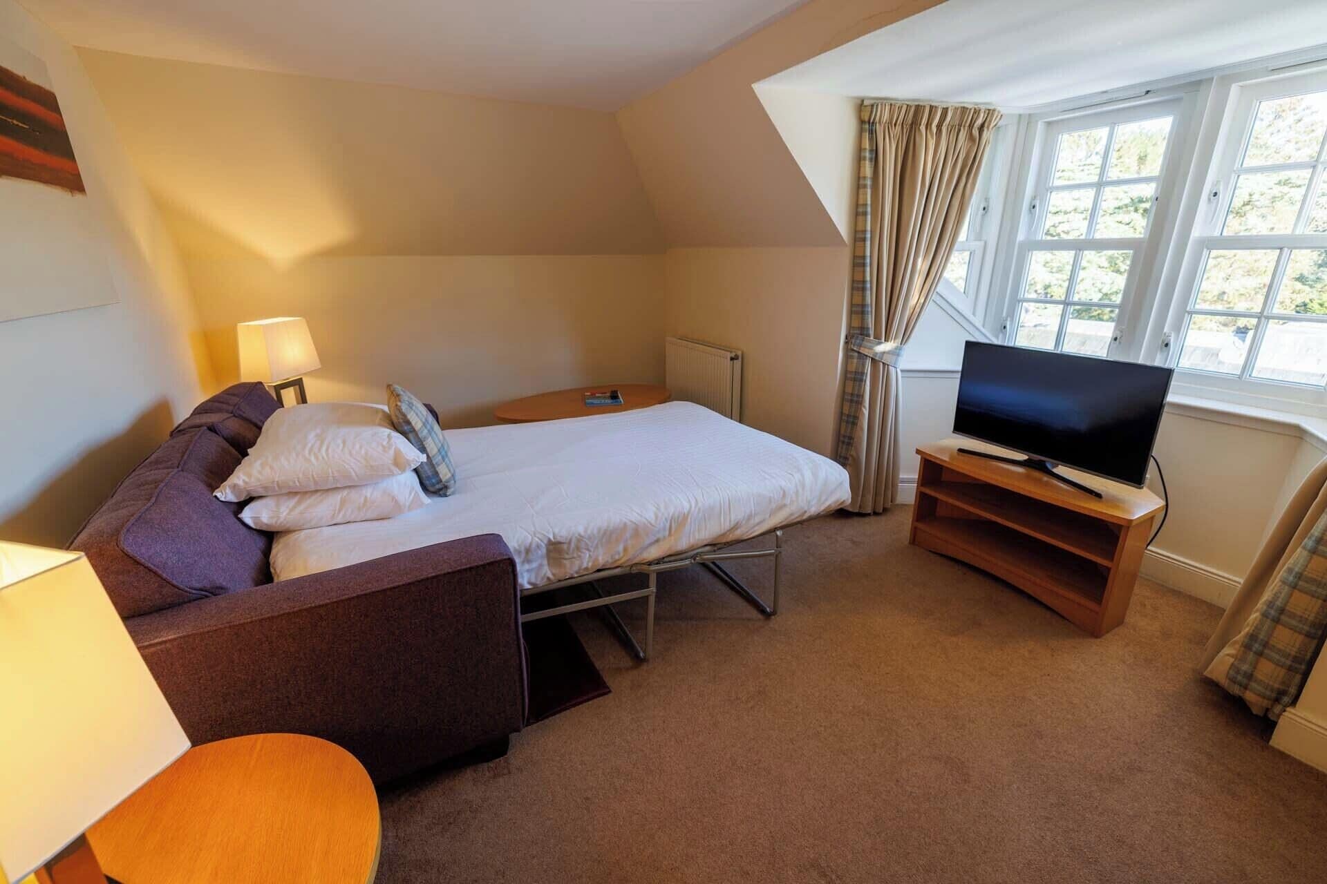 A hotel room with a bed, a TV on a wooden stand, a small table, and a window with curtains.