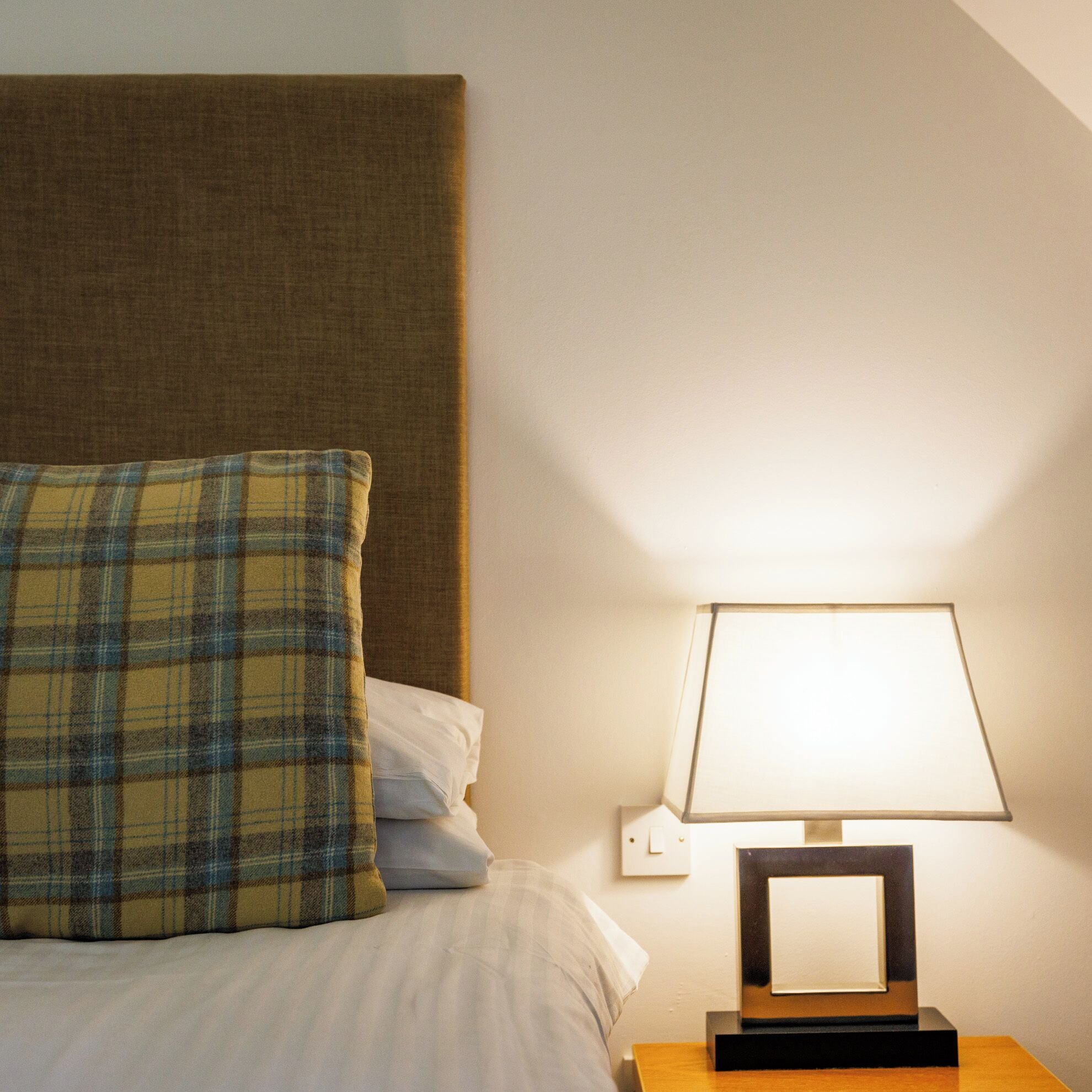 A neatly made bed with a plaid pillow and a bedside table with a lamp.