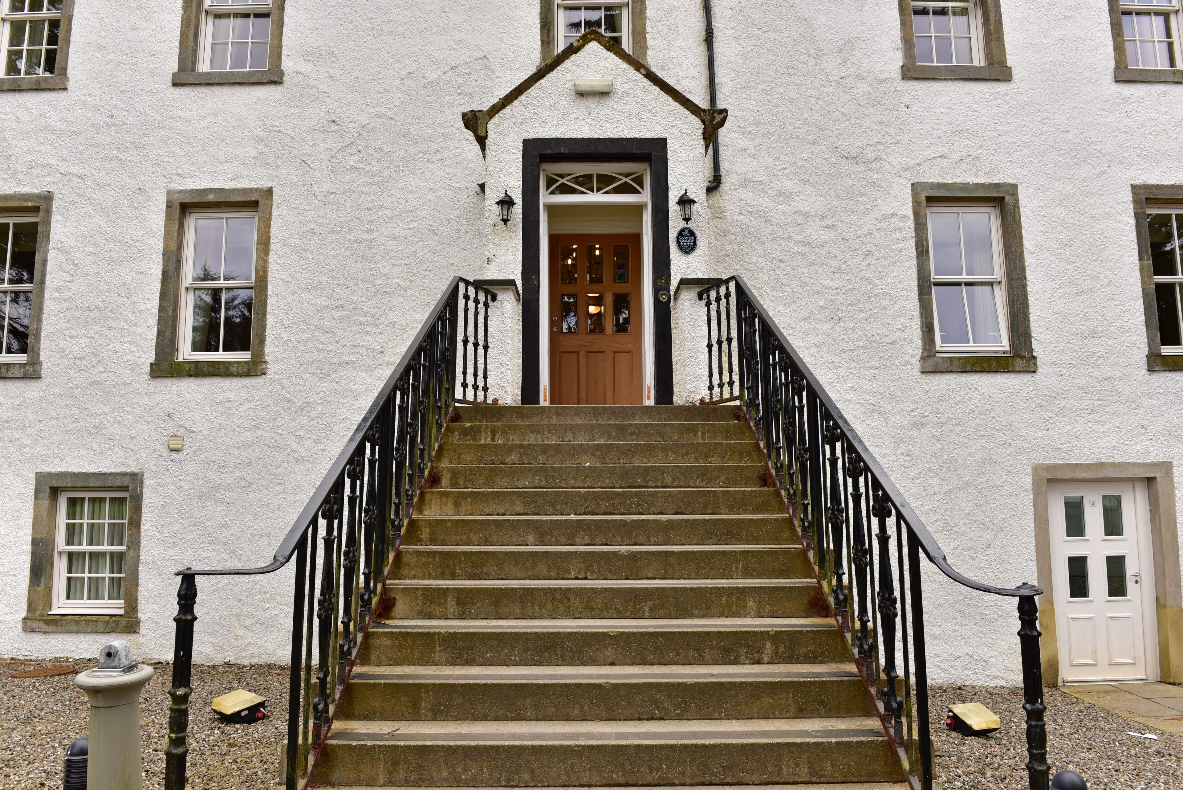Un escalier menant à une porte ornée d’une rampe en fer noir, de part et d’autre de laquelle se trouvent des fenêtres à cadre blanc.