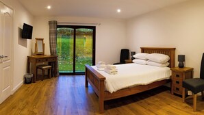 Une chambre avec un lit en bois, un bureau avec un miroir, une chaise, une télévision et une vue sur un jardin, visible à travers une porte vitrée.