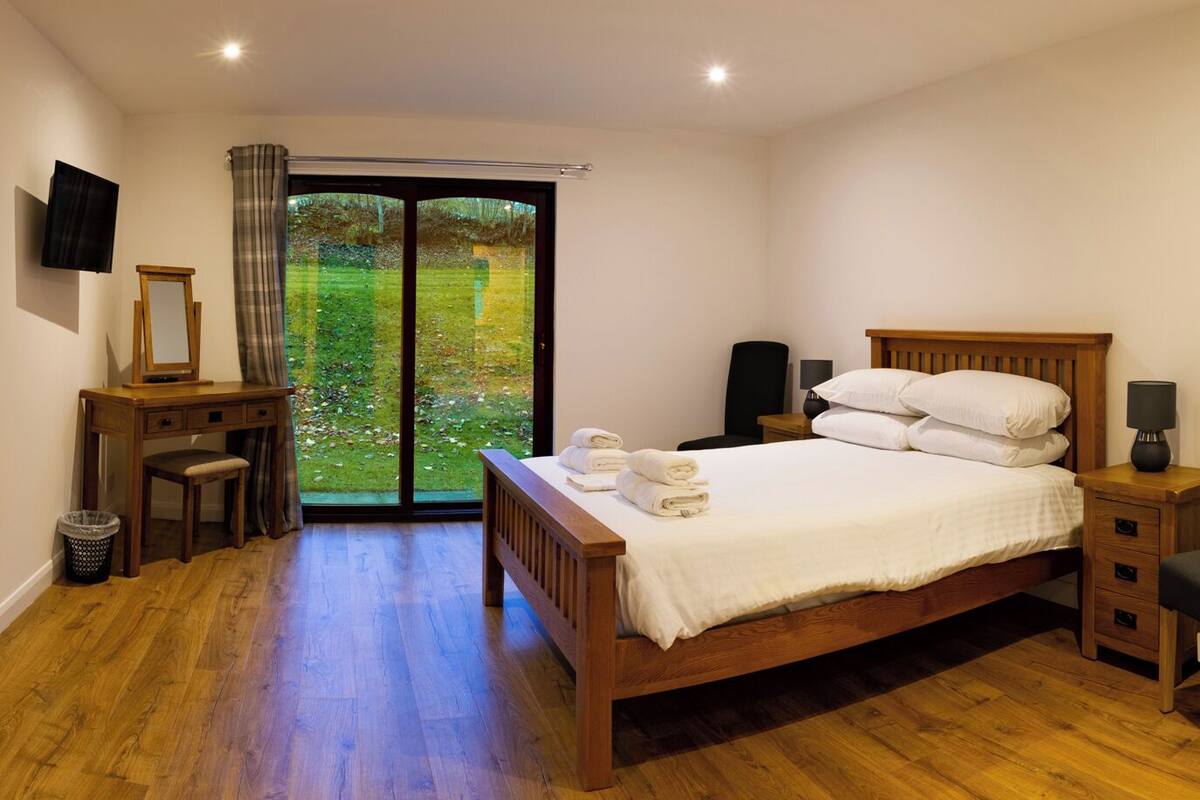A bedroom with a wooden bed, a desk with a mirror, a chair, a TV, and a view of a garden through a glass door.