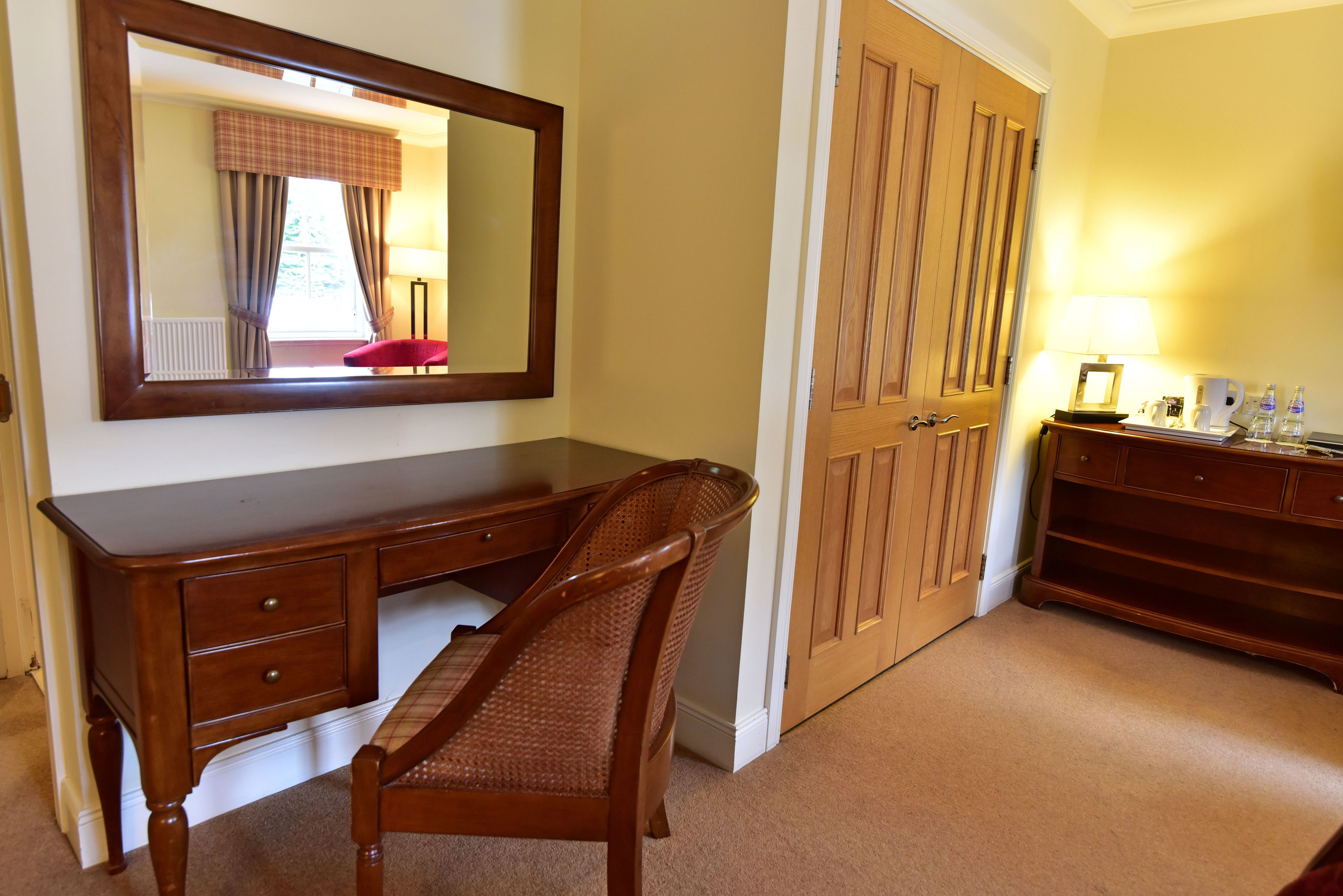 A bedroom with a wooden dresser, a chair, a mirror, a wardrobe, and a bedside table with a lamp and bottles.