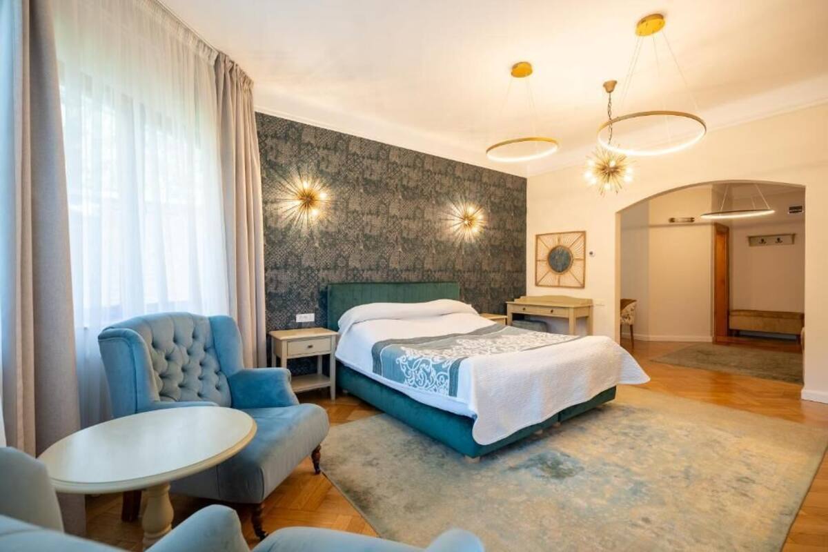 Ein modernes Hotelzimmer mit einem Bett, einem blauen Sessel, einem runden Tisch und einem großen Fenster mit Vorhängen.