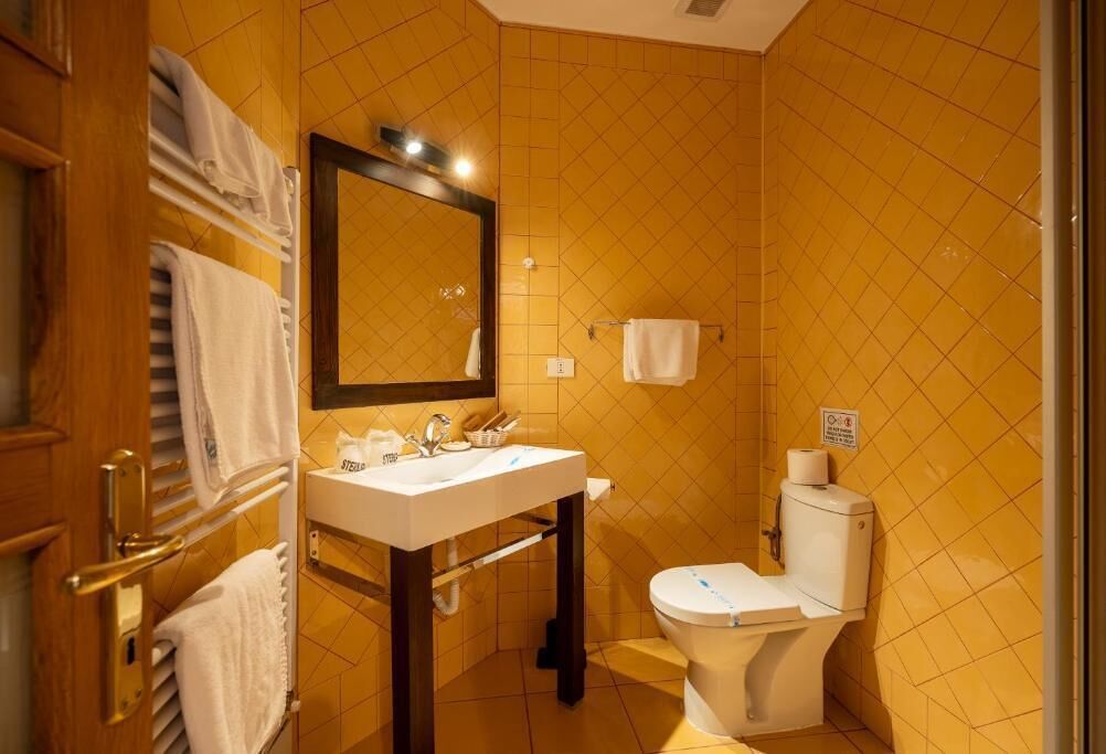 A bathroom with yellow tiled walls, a white sink with a mirror above it, a toilet, and a towel rack with towels.