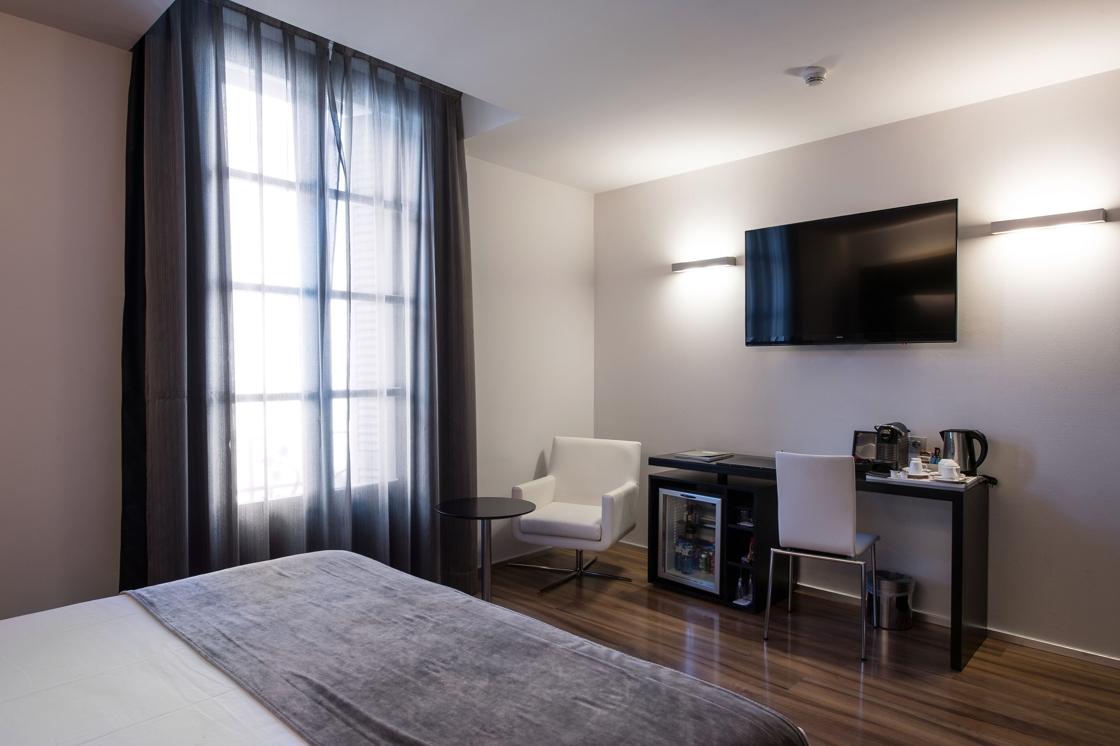A modern hotel room with a large bed, a desk with a computer, a chair, a television mounted on the wall, and a window with curtains.