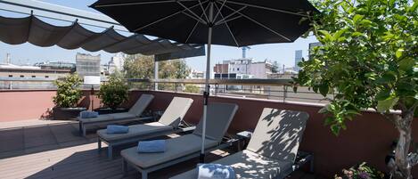 A rooftop terrace with sun loungers, a large umbrella, and potted plants.