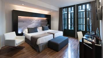 A modern hotel room with a large bed, a sofa, a desk, and a view of the city through large windows.