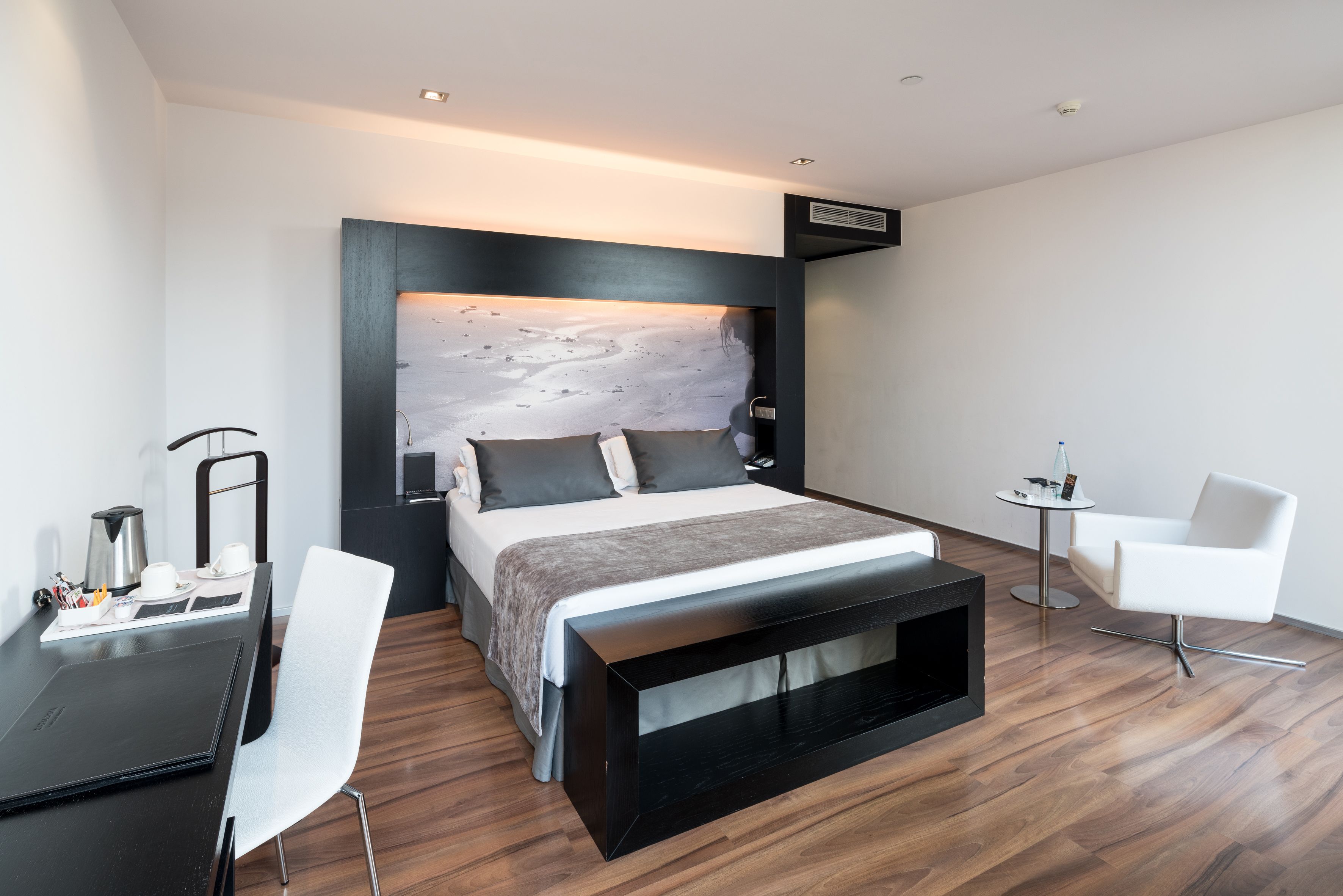 A modern hotel room with a large bed, a small desk, a chair, and a bedside table.
