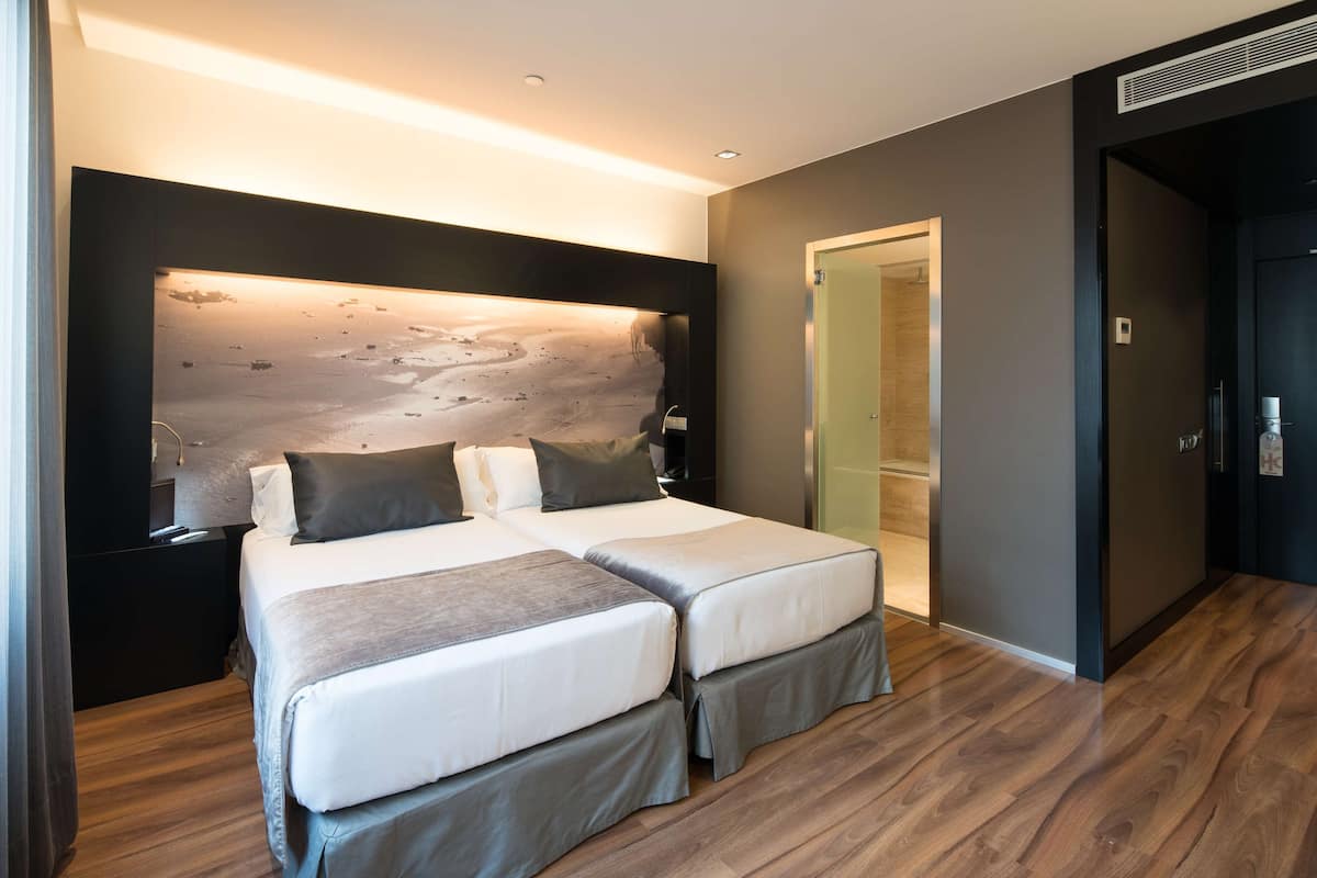 Moderni hotellihuone, jossa on kaksi sänkyä, suuri seinälle asennettu valaisin ja puulattia.