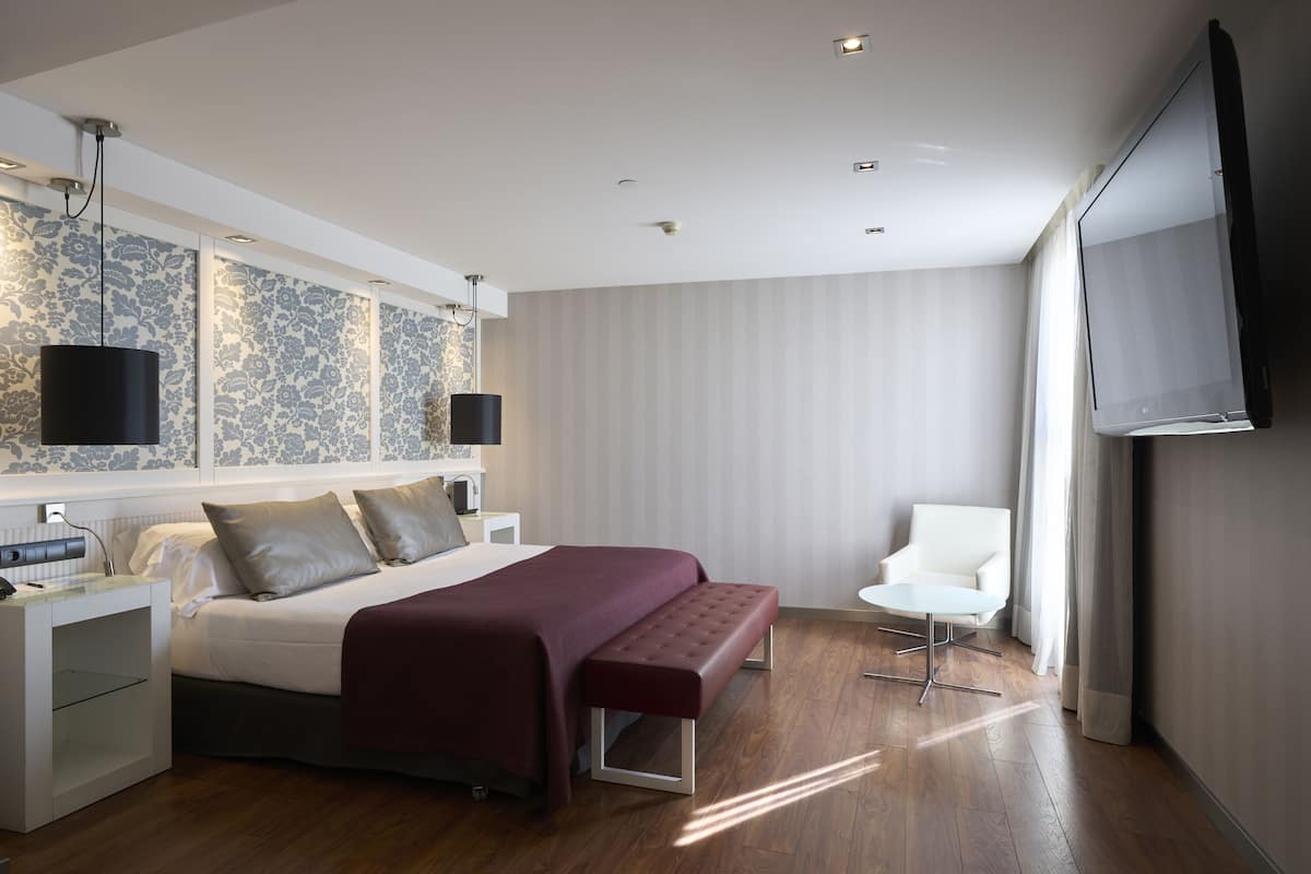 Moderni hotellihuone, jossa on suuri sänky, yöpöytä, penkki, televisio ja tuoli.