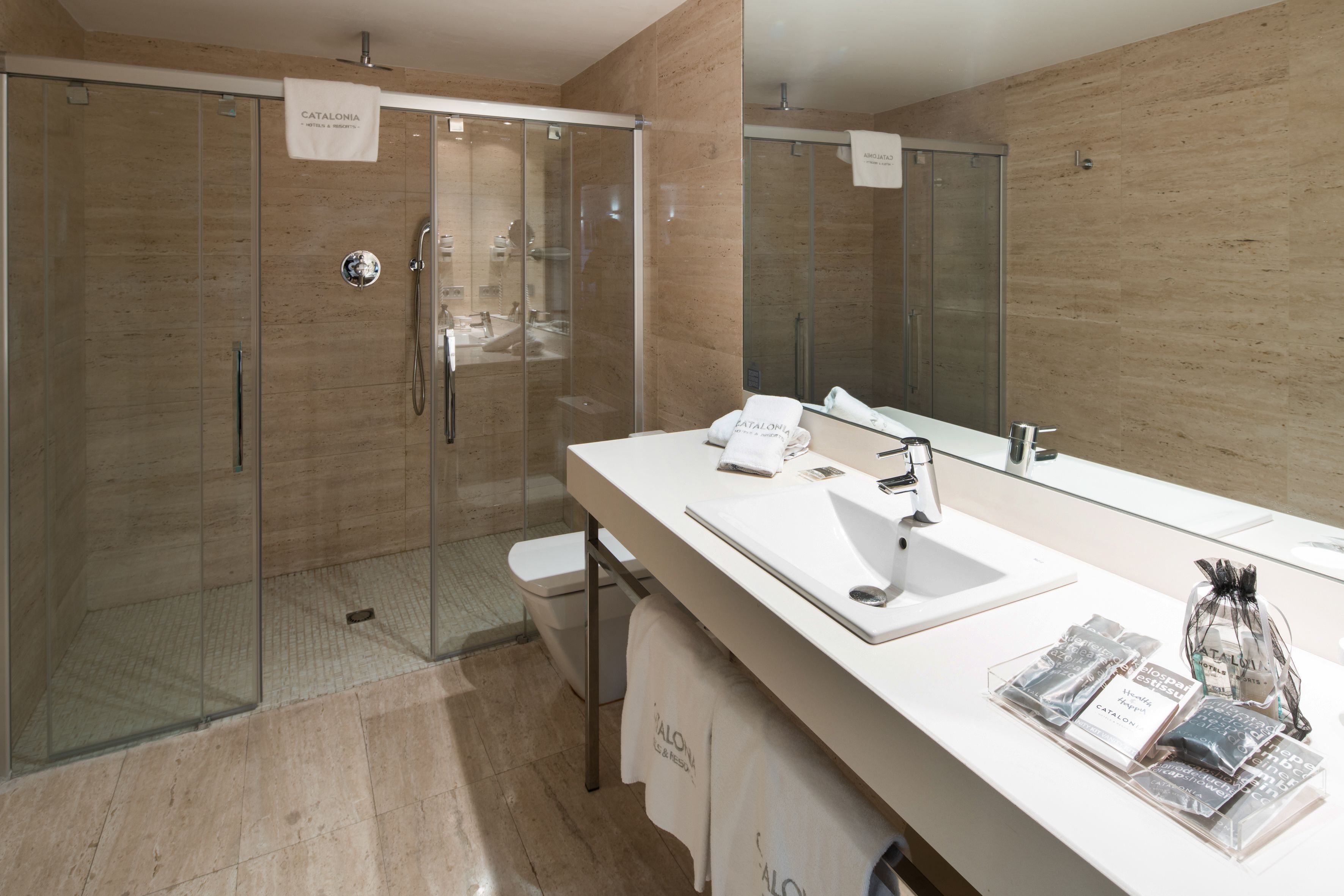 A modern bathroom with a glass-enclosed shower, a large mirror, and a white vanity with toiletries.
