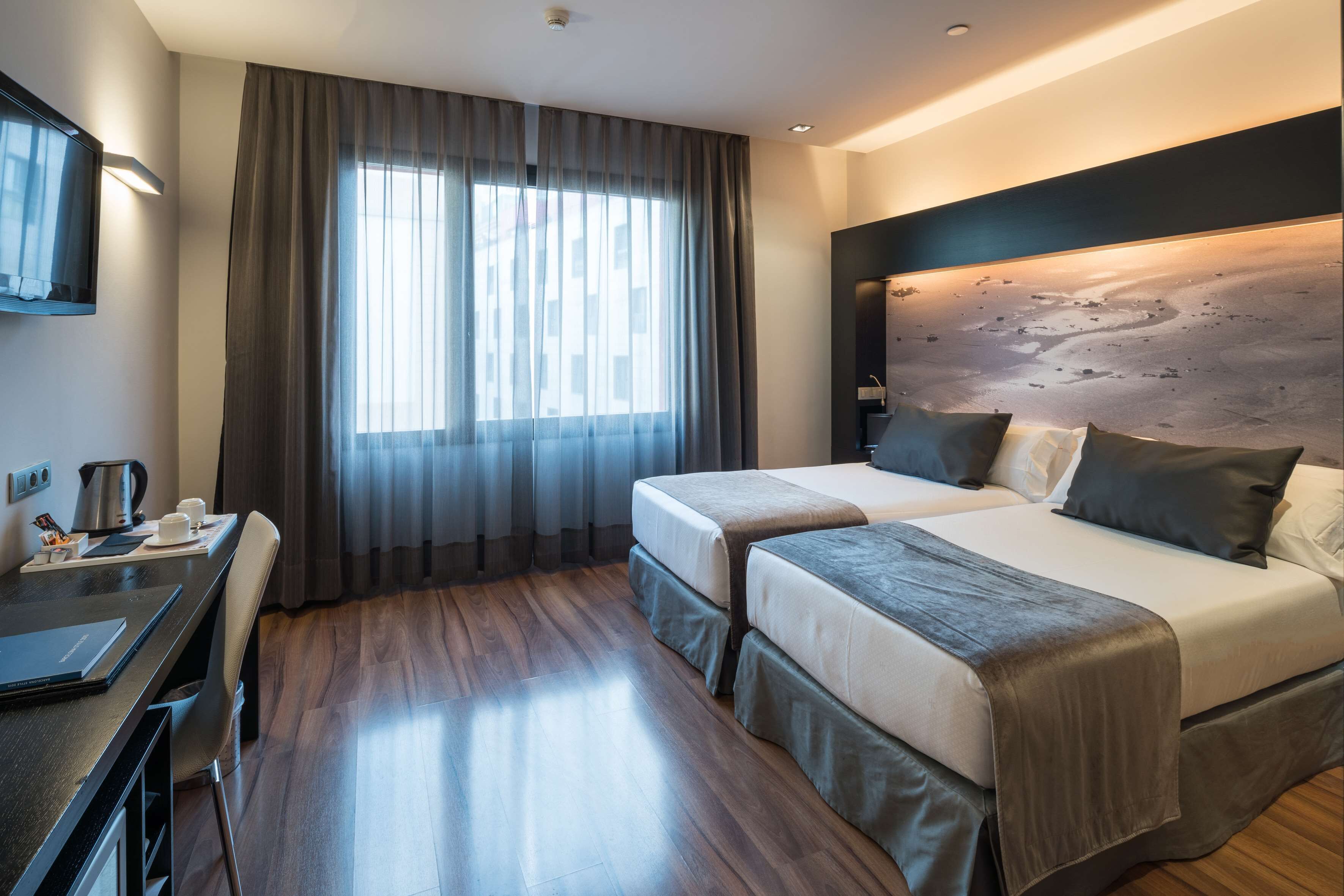A hotel room with two beds, a desk, a chair, a TV, and a large window with curtains.