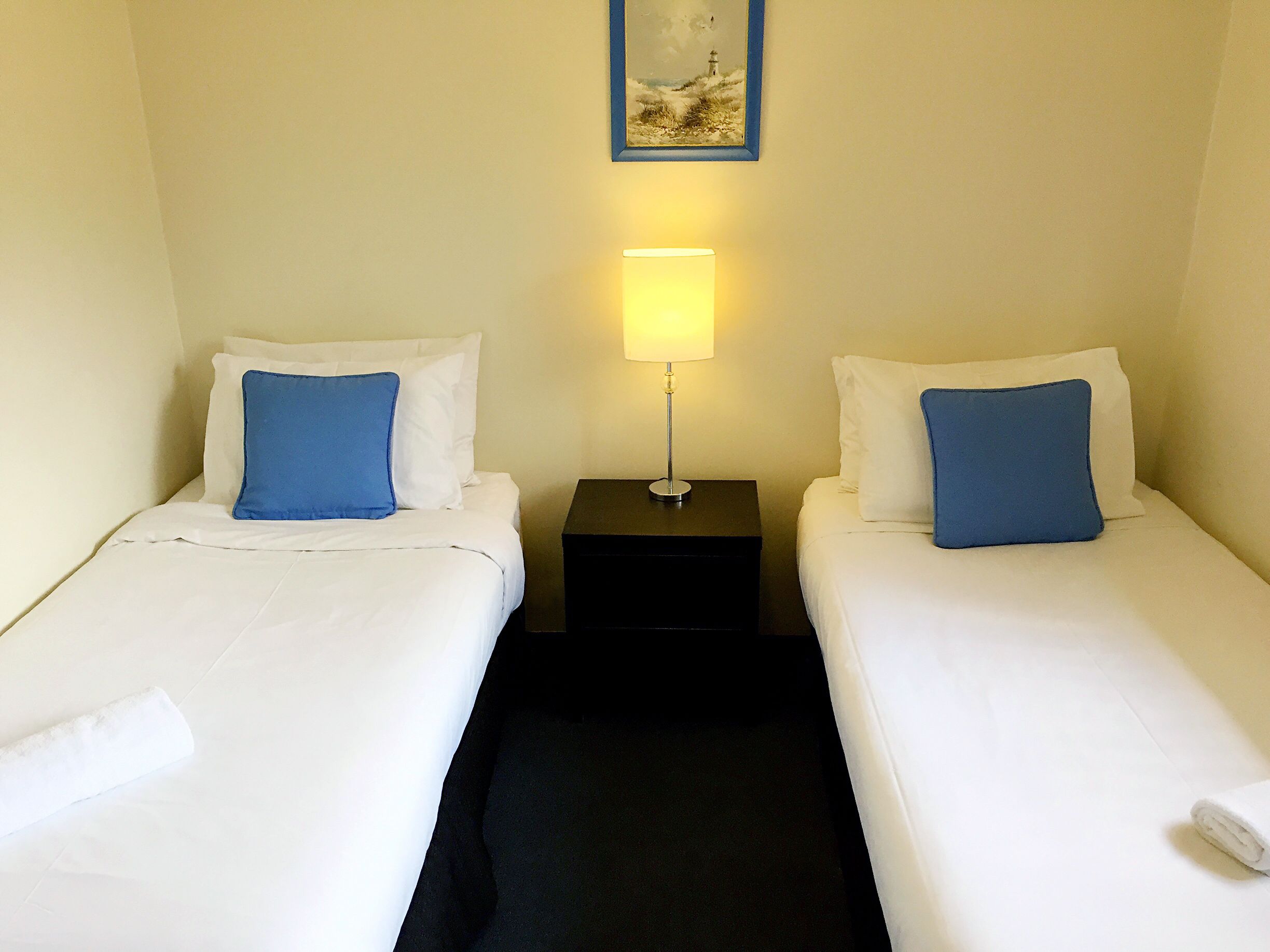 A hotel room with two single beds, each with white bedding and a blue pillow. A nightstand with a lamp is between the beds.