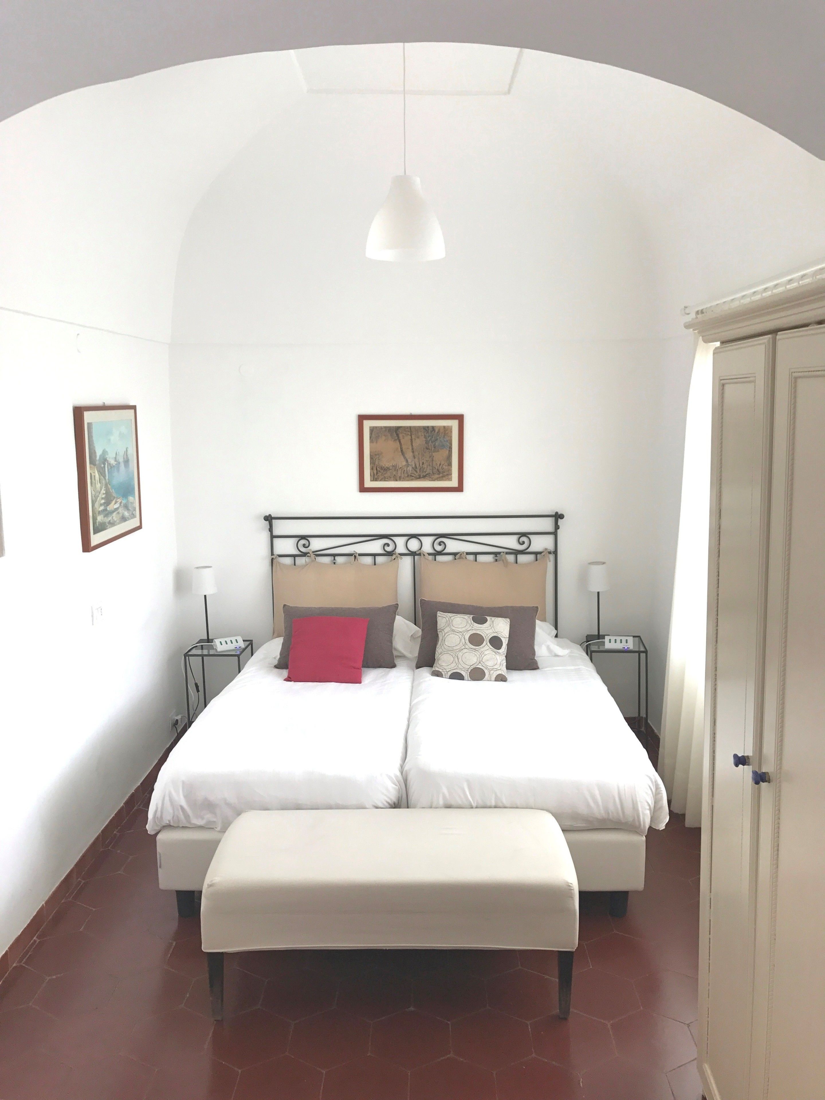 Un lit double avec une banquette, deux tables de chevet, un luminaire suspendu et deux tableaux encadrés au mur.