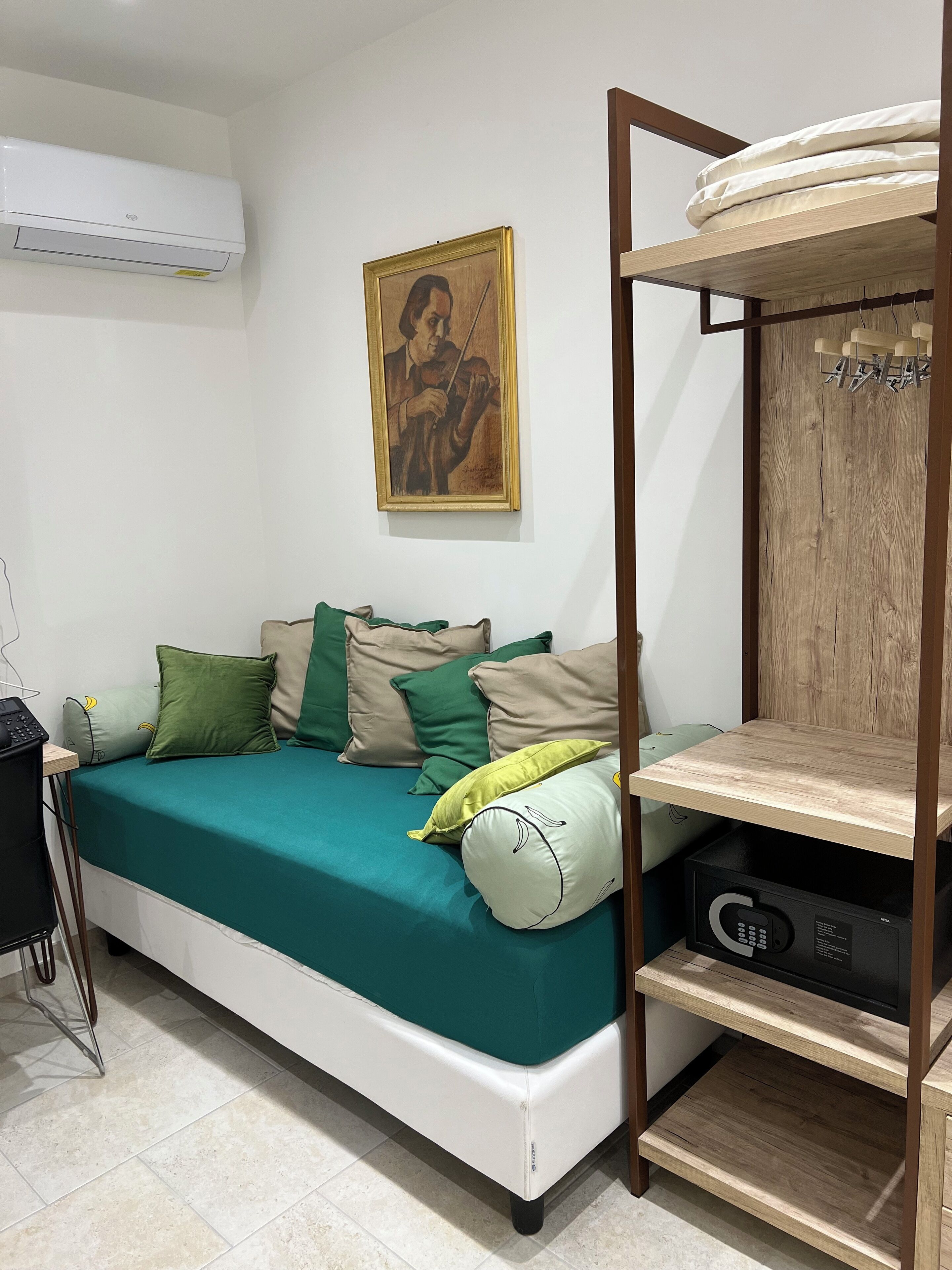 A compact bedroom with a bed, pillows, a wooden shelf, a painting, and an air conditioning unit.