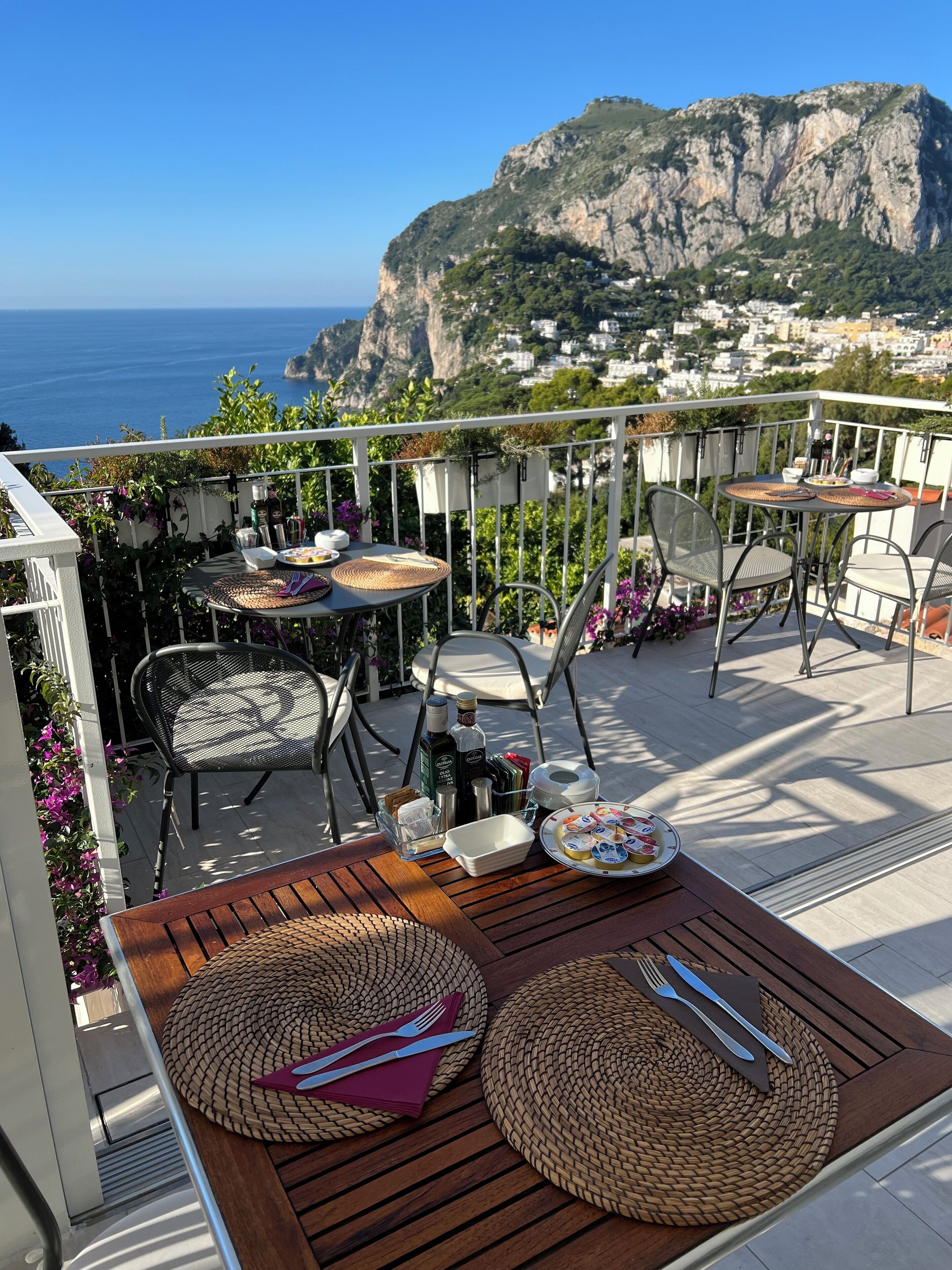 A terrace with outdoor seating, a table set for dining, and a scenic view of a coastal town and cliffs.