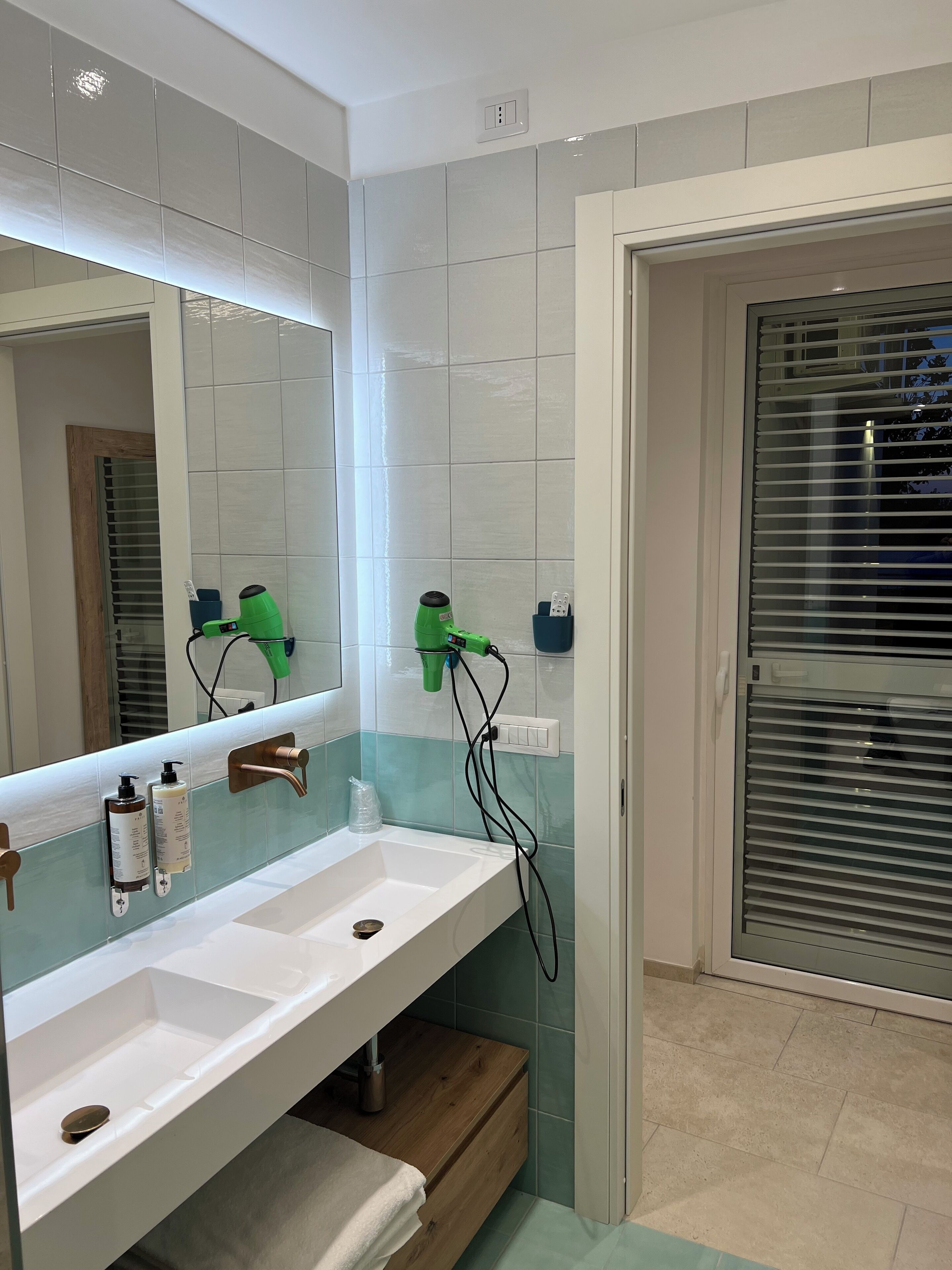 Une salle de bain avec un lavabo blanc, deux bouteilles de shampoing, un sèche-cheveux vert et un miroir.