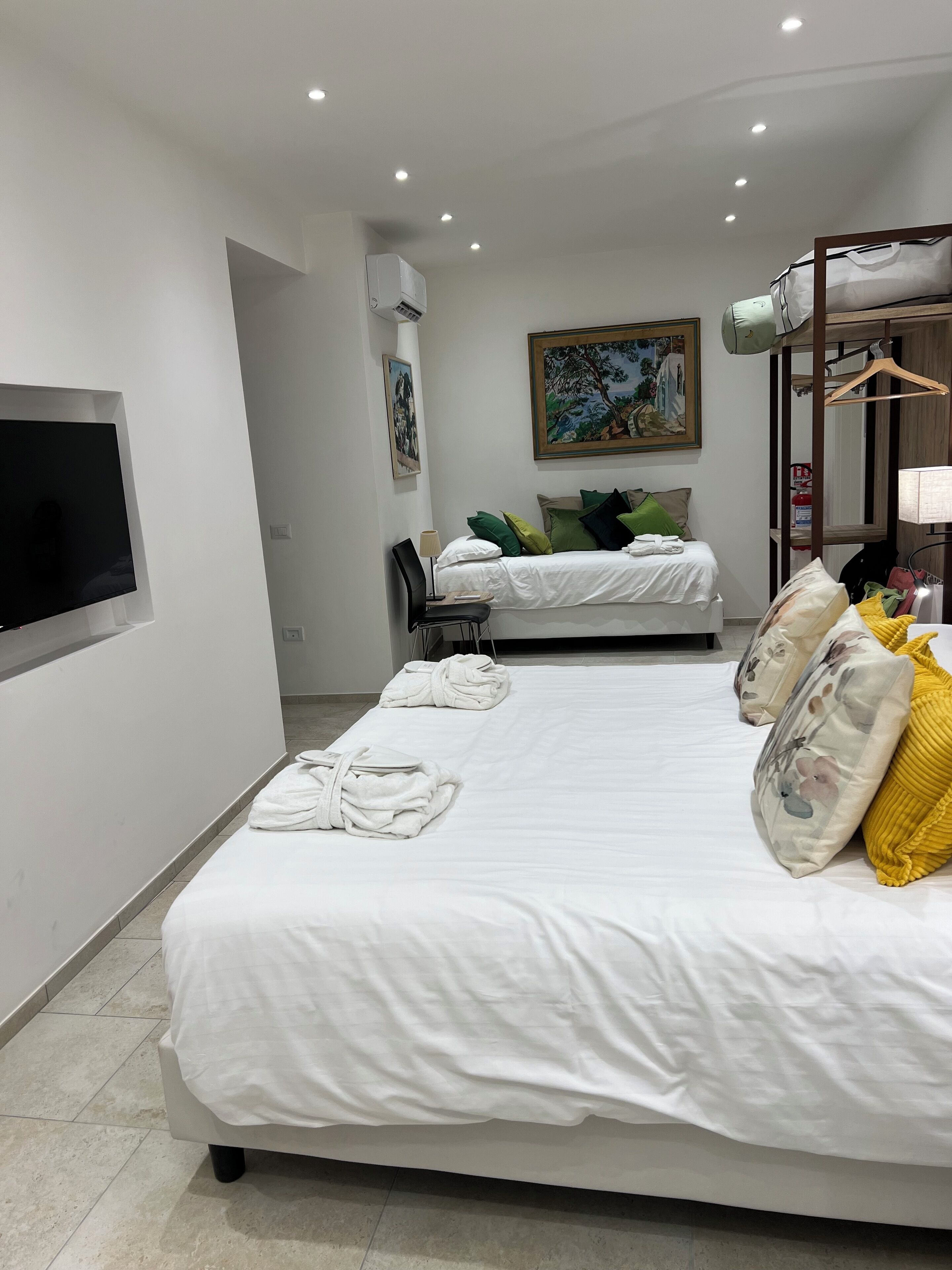 Una habitación de hotel con una cama grande, una cama más pequeña, un televisor montado en la pared, un cuadro y un armario con espejo.