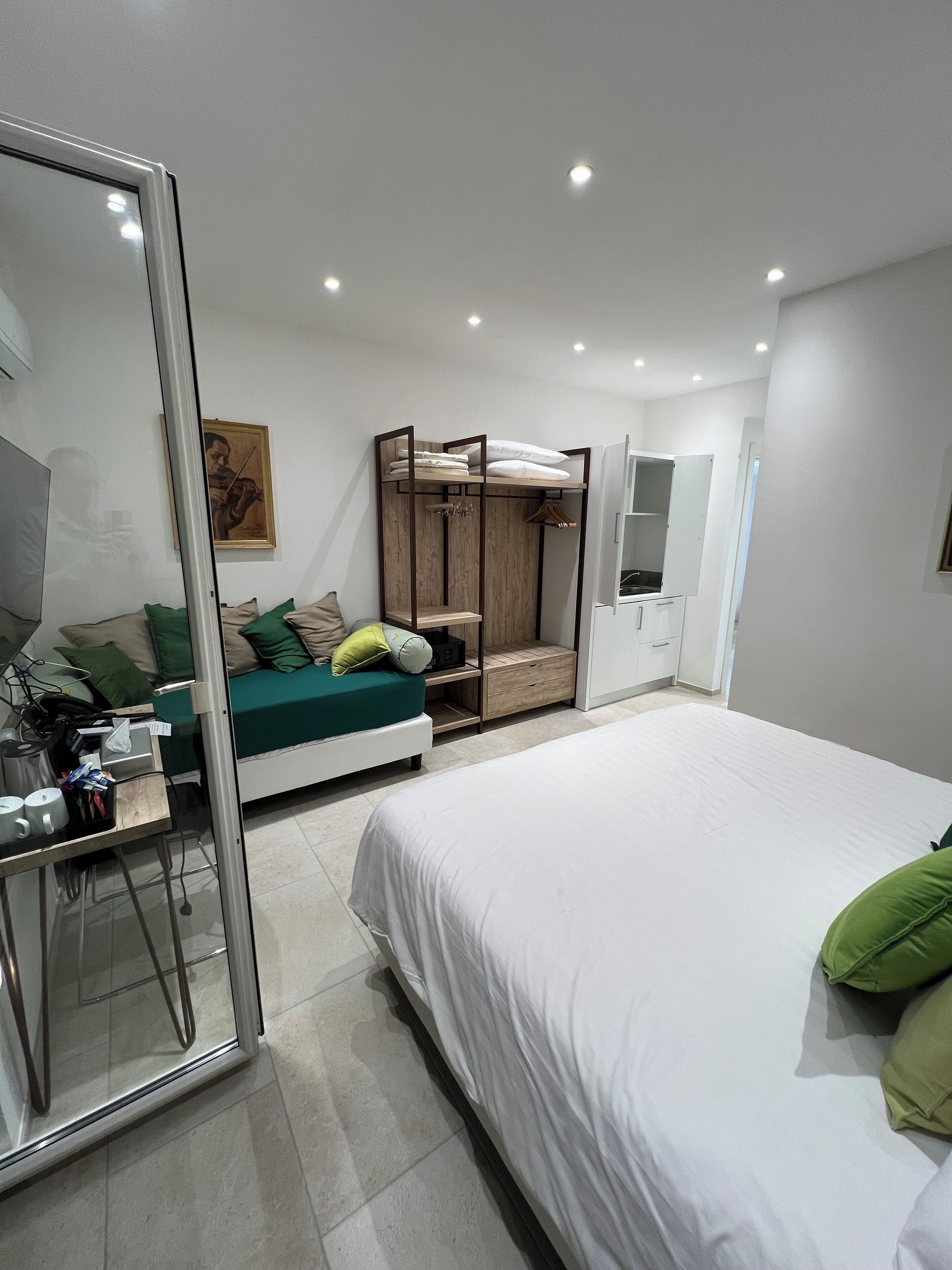 A modern bedroom with a bed, a wardrobe, a seating area with cushions, a mirror, and a painting on the wall.