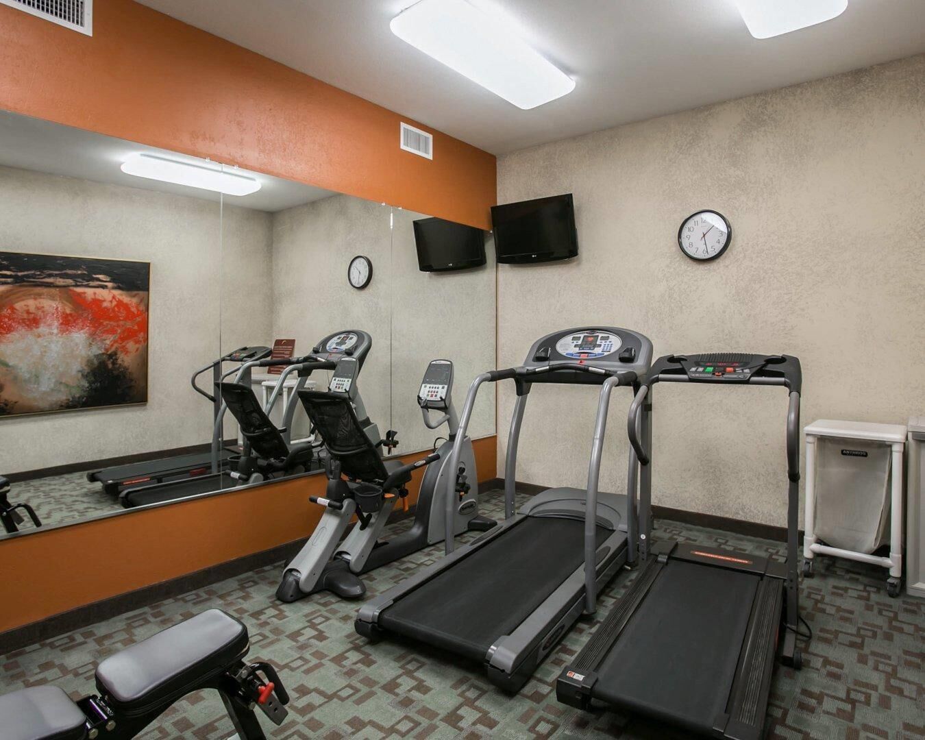 A fitness room with treadmills, an elliptical, a bench, a wall clock, and a television.