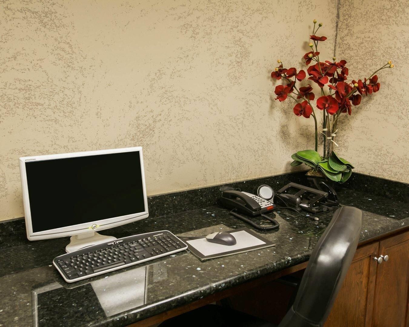 A desktop computer with a keyboard and mouse, a telephone, and a vase with red flowers on a marble countertop.